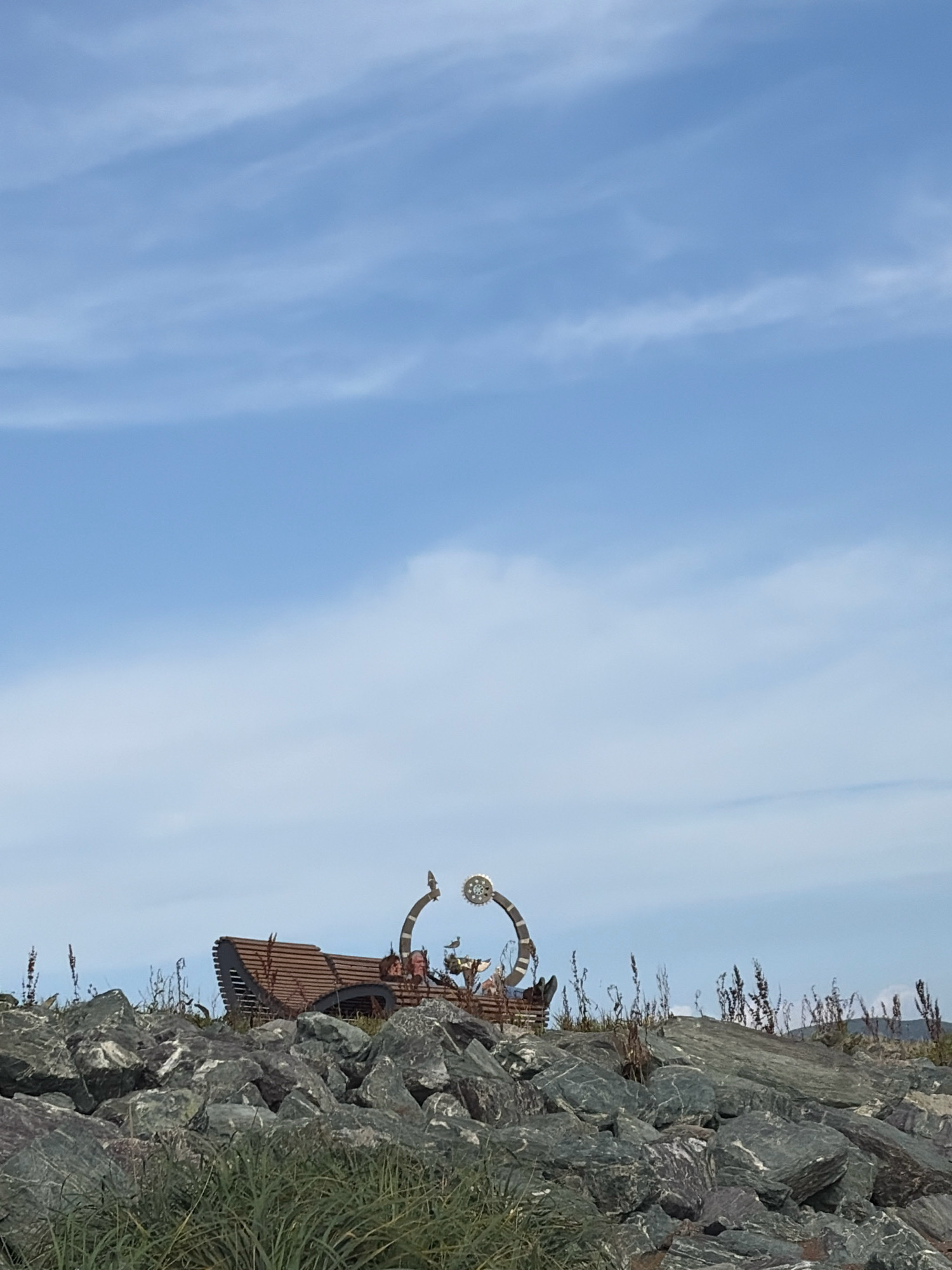 An outdoor scene features a large, gray, rocky embankment in the foreground, with green grasses and dry stalks of vegetation growing between the rocks. Atop the embankment, near the center of the frame, is a unique wooden bench with a curved, slatted backrest. Behind the bench, a circular metal sculpture with intricate gears and decorative elements is visible, arching upwards and to the right. The sculpture appears to be a stylized clock face or celestial object. A few small figures or decorative elements can be seen on the bench and near the sculpture, including what looks like a bird. The background consists of a vast expanse of blue sky with wispy white clouds. The overall setting suggests a public space or park area, possibly with a scenic view overlooking water, given the embankment and open sky. The presence of the bench and sculpture implies a decorative or commemorative purpose for the location.