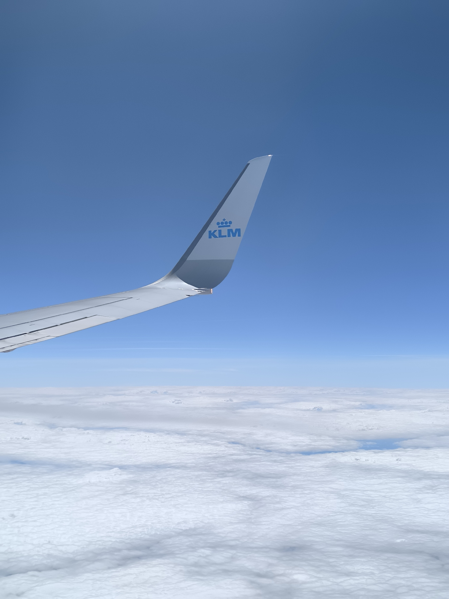 A view from an airplane window shows a KLM airplane wing against a backdrop of blue sky and a blanket of clouds. The KLM logo is visible on the wing. The location is not in Fischamend, Austria.