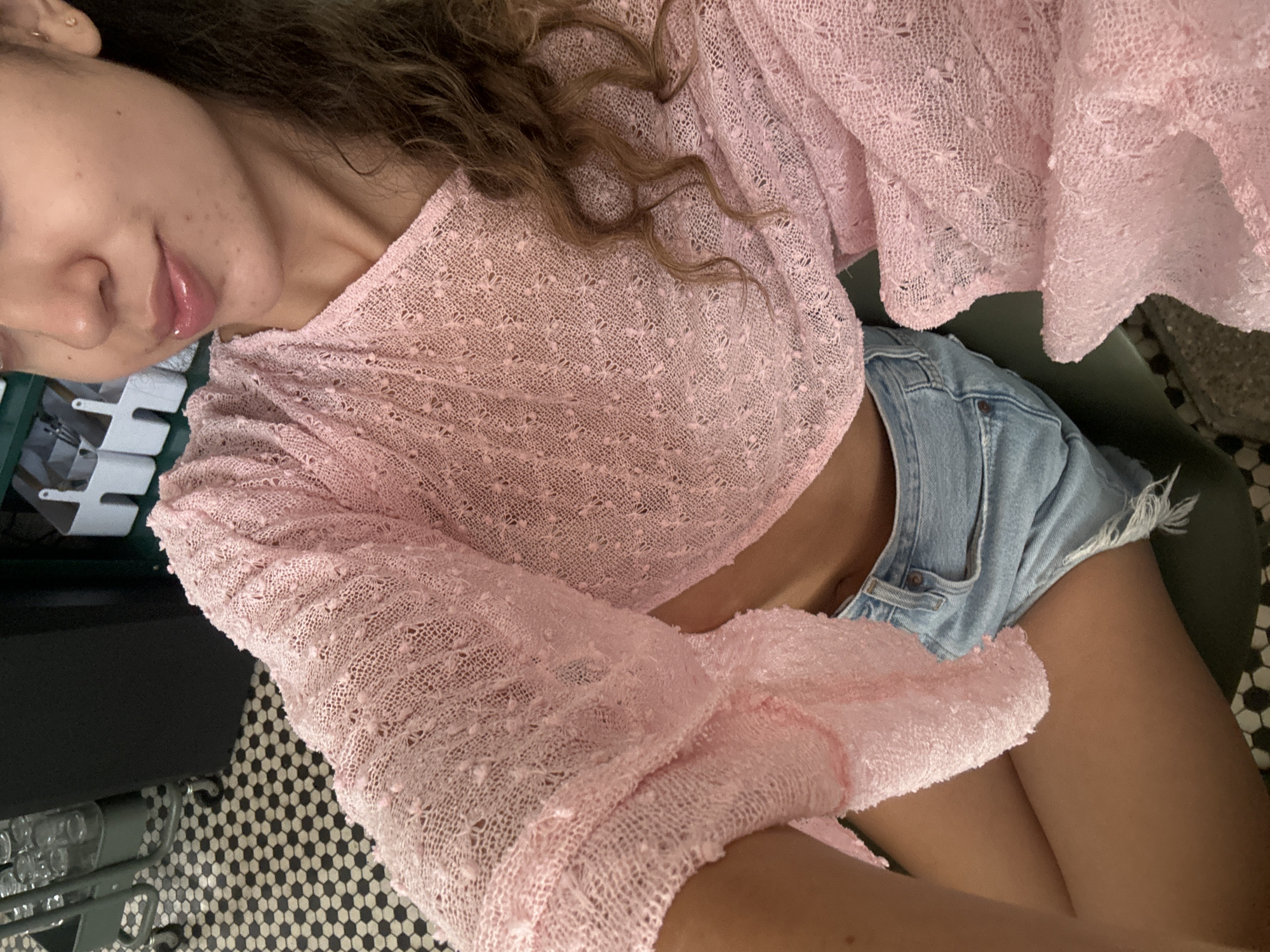 The image shows a close-up view of a young woman wearing a pink, loosely knit, cropped top with bell sleeves and light-wash denim shorts. She is sitting, with her legs crossed and her upper body angled towards the camera. Her hair, which is wavy and brown, falls over her shoulder and onto the pink top. Her lips are glossed and slightly parted. The background is blurred but appears to be an indoor setting with a patterned floor, possibly tile, and some shelving units with white containers. There are no recognizable landmarks or details that would indicate a specific location within San Juan, United States of America. The lighting is soft and appears to be natural or from an overhead source.