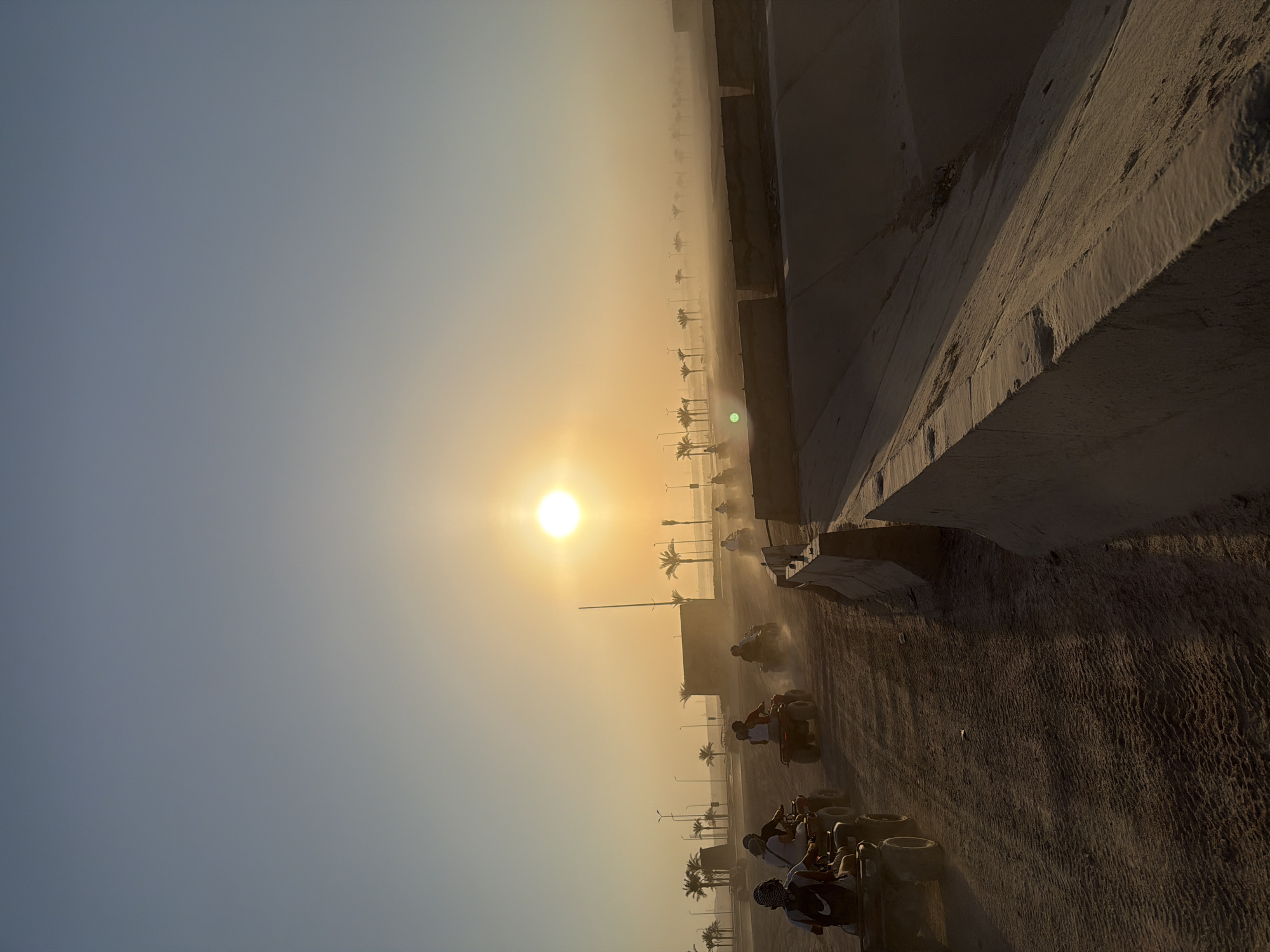The image depicts a group of people riding ATVs in Sharm El Sheikh, Egypt, during sunset. The sun, a bright orb, hangs low in the sky, casting a warm, golden light that creates a hazy atmosphere. In the foreground, several ATVs are kicking up dust as they traverse a sandy path. The riders, silhouetted against the sun, appear to be enjoying an adventurous activity. Palm trees dot the landscape, characteristic of the region. The scene suggests a tourist activity, likely a desert safari, taking place in a coastal desert environment. The presence of concrete barriers and a visible construction in the background may indicate an area undergoing development or a designated tourist zone. The overall impression is one of adventure and natural beauty, with the sunset adding a dramatic and picturesque quality to the scene.