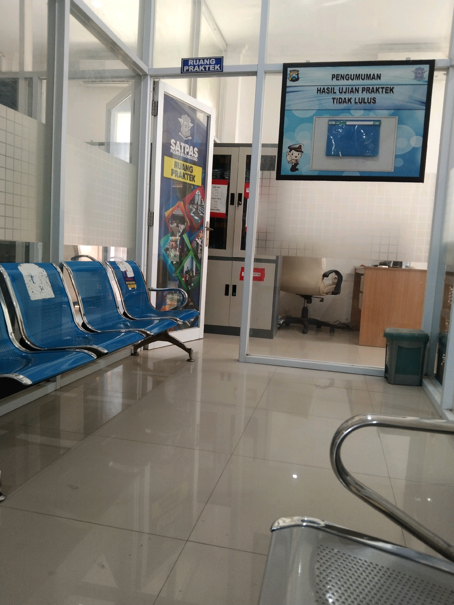 The image depicts an indoor waiting area at what appears to be a public service office, possibly related to driver's license testing, given the presence of signs in Indonesian. The room is furnished with several rows of blue, metal waiting chairs with chrome legs. The floor is tiled with large, light-colored, reflective tiles.
On the left side of the image, a frosted glass partition separates the waiting area from another room. A sign above the entrance to this room reads "RUANG PRAKTEK" (Practice Room). To the right of this entrance is a door with a large graphic display that includes text such as "SATPAS POLRES BOJONEGORO" (Motor Vehicle Registration Center, Bojonegoro Police).
On the right side of the waiting area, there is a framed announcement board with the text "PENGUMUMAN HASIL UJIAN PRAKTEK TIDAK LULUS" (Announcement of Practice Test Results Not Passed). Below this text is an image of a computer screen and a small cartoon figure of a person in a hat. Further to the right, a glass partition reveals an office space with a desk, an office chair, and a wooden cabinet. A green trash can is visible in the foreground on the right.
The environment is clean and well-lit, with the overall impression of a functional, administrative space. The presence of the "Bojonegoro" text on a sign strongly suggests the location is within the city of Bojonegoro, Indonesia. There are no people clearly visible in the waiting area itself, though a person's face is reflected in the glass partition in the office space.