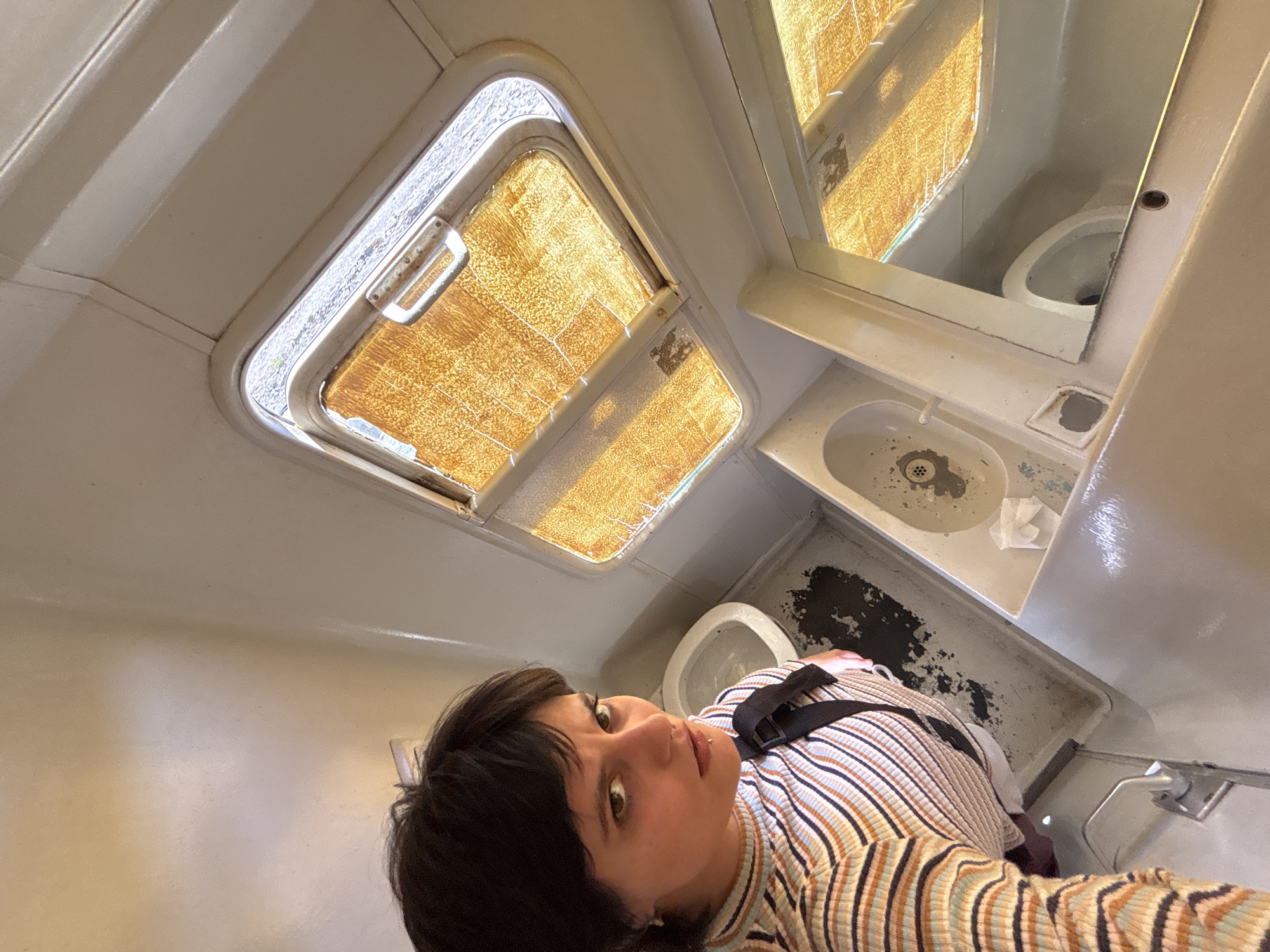 The image is a high-angle selfie taken inside a small, utilitarian restroom, likely on public transport in Sofia, Bulgaria. The central subject is a person with short, dark hair and dark eyes, looking directly at the camera. They are wearing a striped long-sleeve shirt with beige, brown, and light blue vertical stripes, and a black strap is visible over their shoulder.
The cramped setting features off-white, aged walls and fixtures. Above and behind the person, a small, rectangular window with a textured or frosted pane allows bright natural light to illuminate the space, suggesting it is daytime. The window frame appears worn, and a handle is visible on the left side.
To the right of the window, a mirror reflects part of a white toilet bowl. Below the mirror is a small, off-white sink with a metal drain cover showing some grime or debris, and a small piece of white paper (possibly tissue) resting on its edge. The floor or base surrounding the sink and toilet exhibits noticeable dark stains or grime, indicating significant wear and lack of pristine cleanliness. The overall aesthetic suggests an older, heavily used facility, consistent with public transportation like a train or bus. No discernible text is visible within the frame.