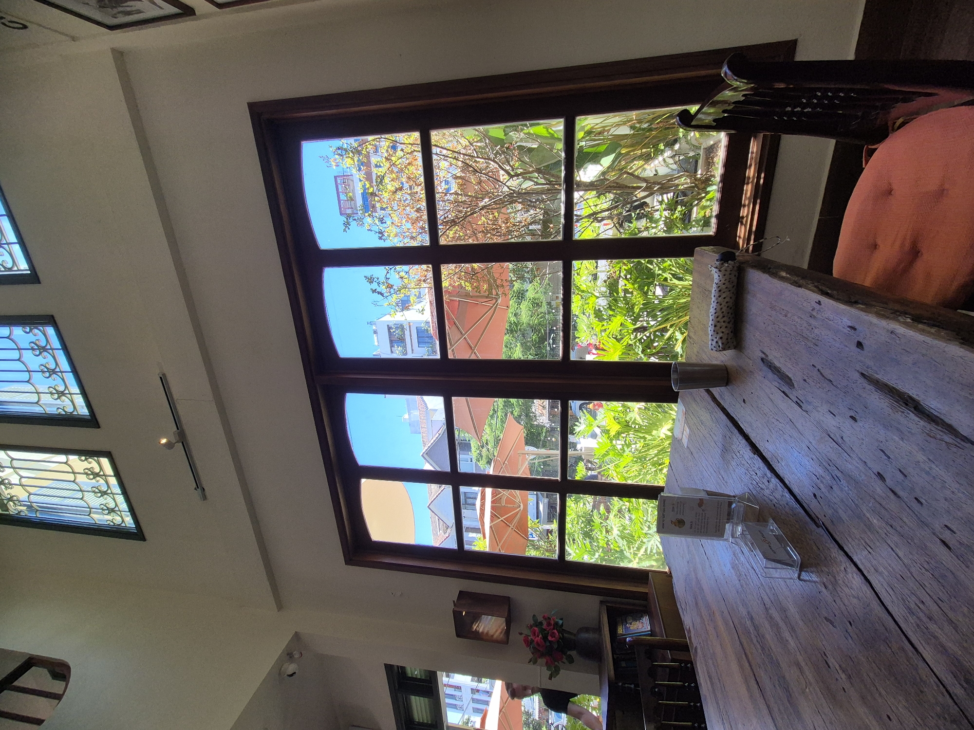 The image depicts the interior of a cafe in Da Nang, Vietnam, with a large window overlooking a lush outdoor area. A weathered wooden table occupies the foreground, set with a glass of water, a small umbrella, and two clear acrylic stands holding menus or cards. To the right, a dark wooden chair with an orange cushion is partially visible. The window is divided into multiple panes, revealing a vibrant scene outside. Visible through the window are orange umbrellas, dense green foliage, and buildings in the background. A man in a dark shirt is seen through a smaller adjacent window to the left, standing near a bookshelf with a vase of red roses. The cafe's interior features high ceilings and decorative elements in the upper windows.