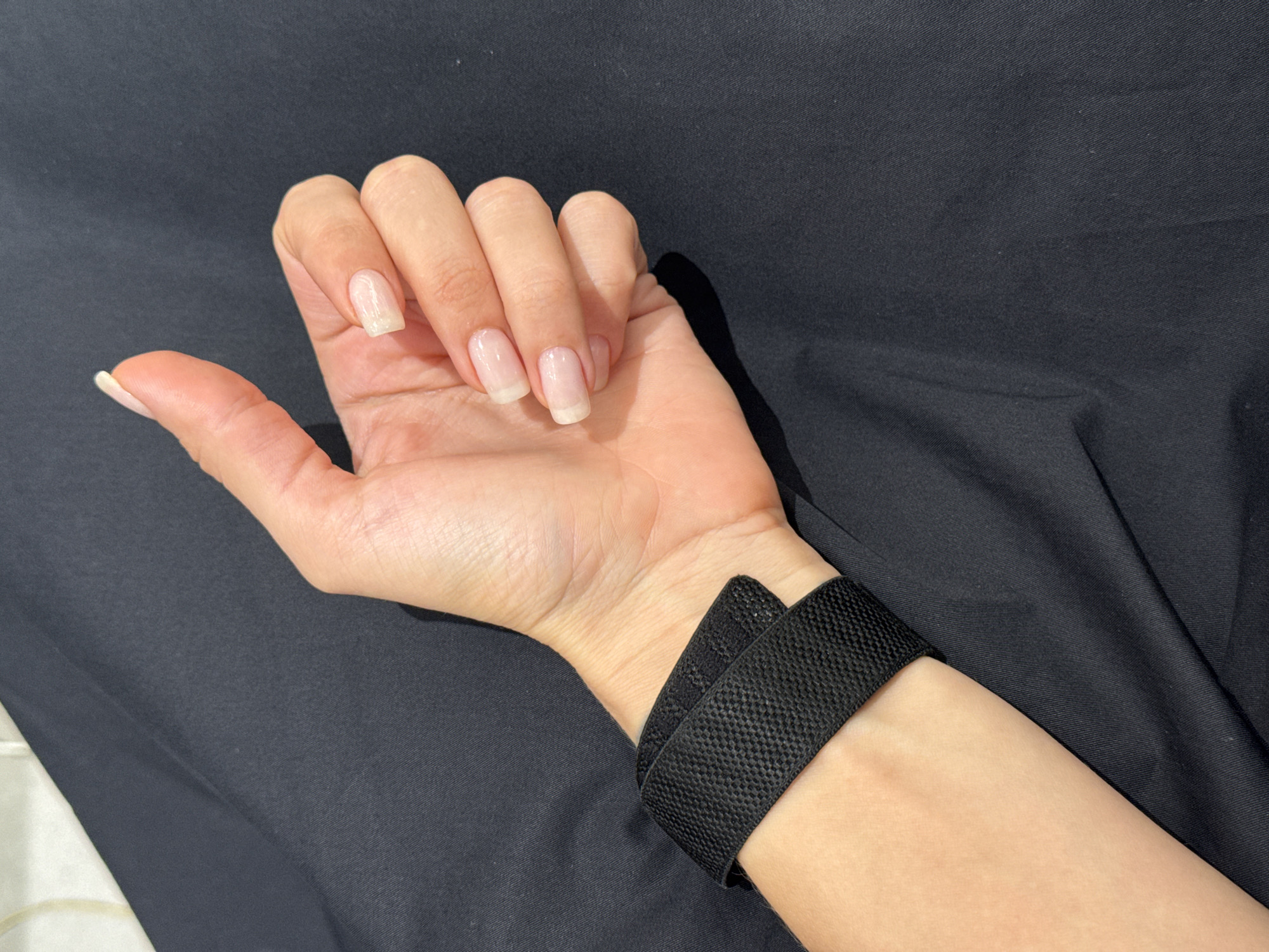 A fair-skinned woman's hand rests on a black cloth background. The woman has a black wrist strap around her left wrist. Her nails are medium length and manicured with a French tip style. The location is not specified, and no other details regarding the city of Dubai or the United Arab Emirates are discernible from the image.