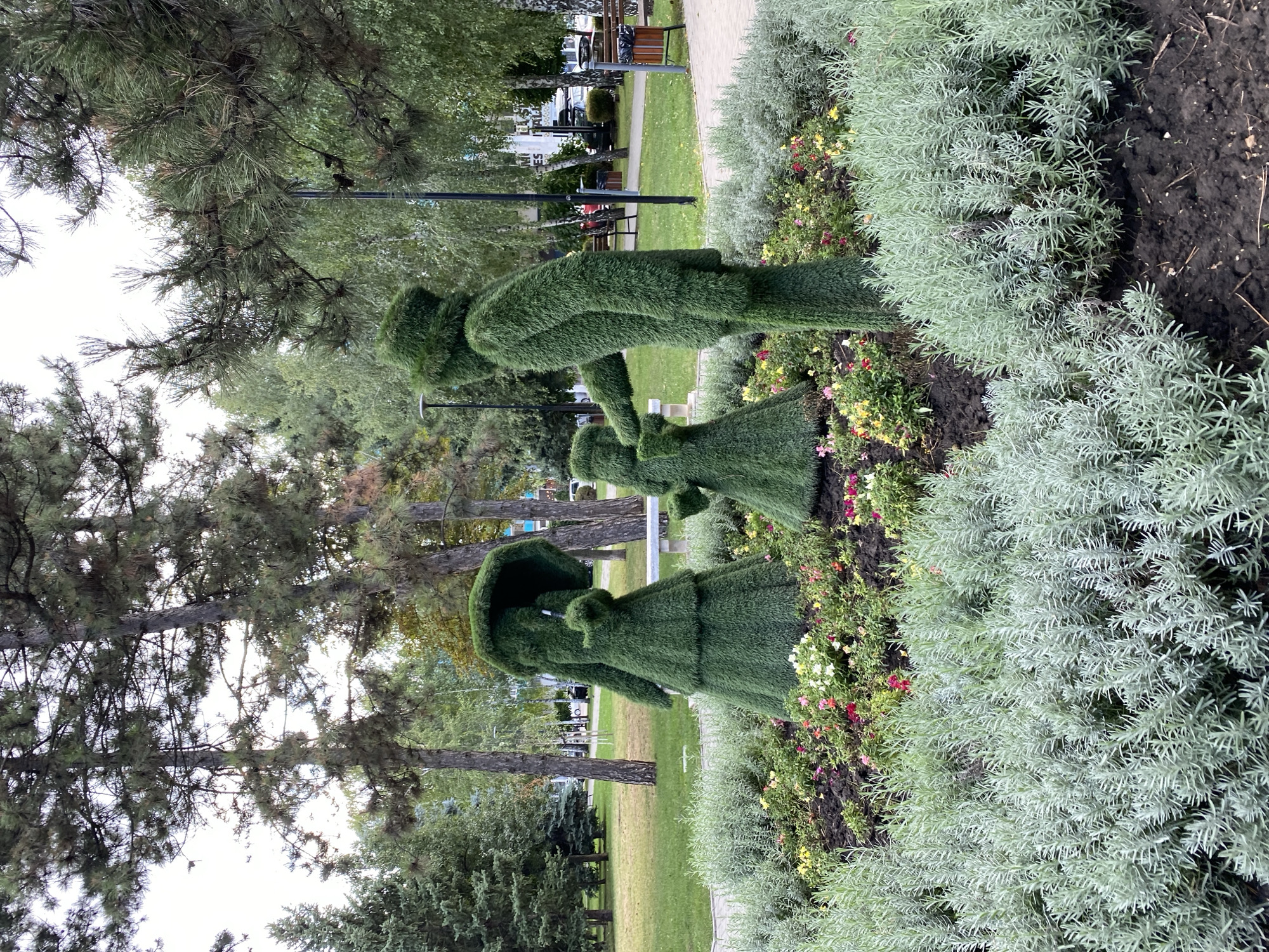 The image depicts a topiary sculpture of a family in a park setting. A tall, slender figure, presumably the father, stands on the right, with a smaller figure, likely a child, in front of him. To the left, a larger figure, possibly the mother, holds an umbrella over both smaller figures. The topiaries are made of lush green foliage and are arranged in a flowerbed with colorful flowers and light green, shrub-like plants in the foreground. In the background, trees, a paved walkway, benches, streetlights, and buildings suggest an urban environment. The sky is overcast. The topiary sculptures appear to be located in a public park in Chișinău, Moldova, given the context of the provided image. The scene captures a moment of family togetherness, with the father's hand outstretched towards the child, and the mother providing shelter. The overall impression is one of a charming and well-maintained public space.