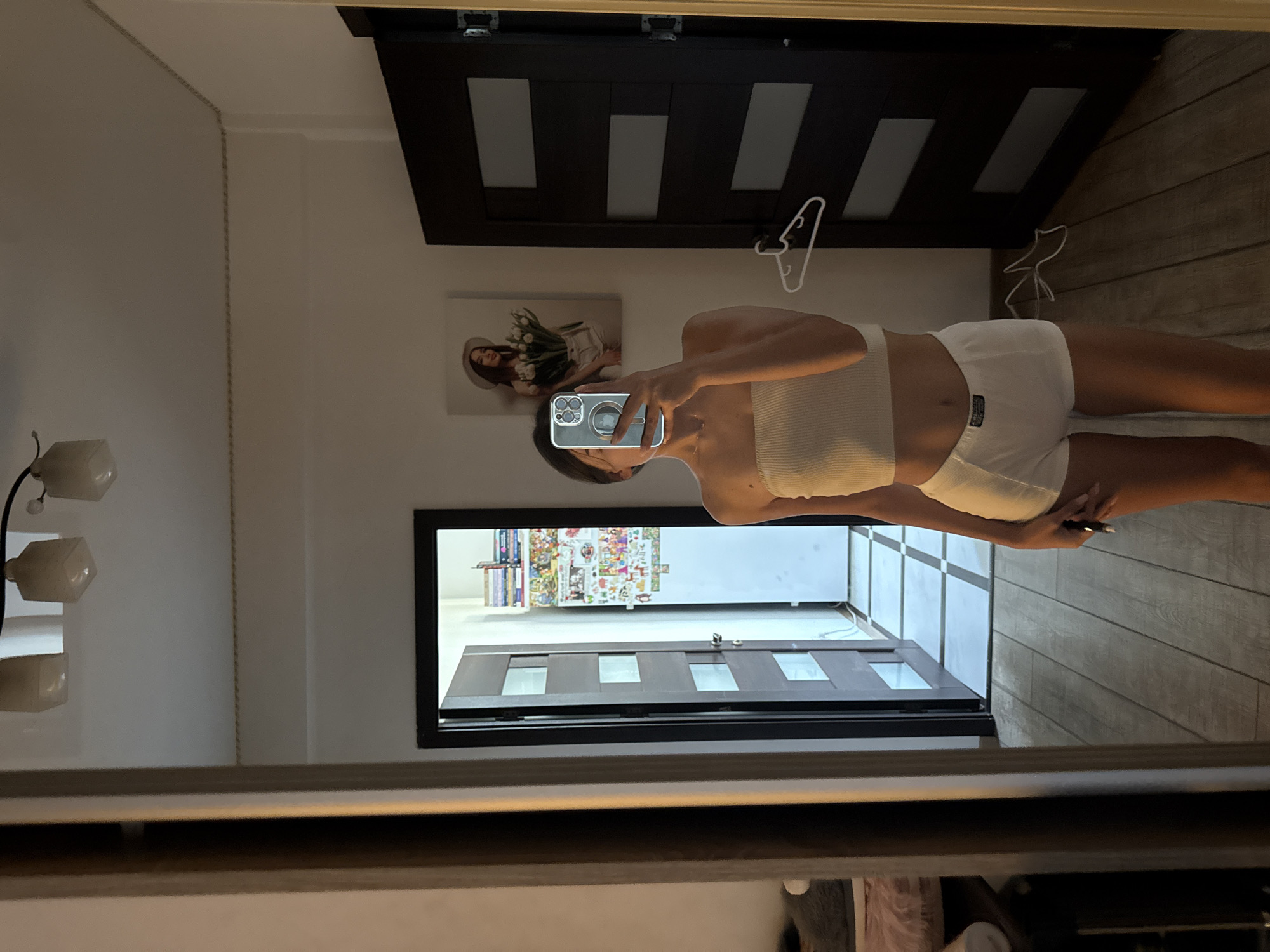 The image shows a woman taking a mirror selfie in what appears to be a bedroom or dressing area. She is wearing a white tube top and white shorts. A phone is held up to take the picture. In the background, there is a closed door with windows and a door frame with a hanger on it. There is also a framed picture on the wall depicting a woman holding flowers. The floor is made of wooden planks. The location is not specified and cannot be determined from the image.