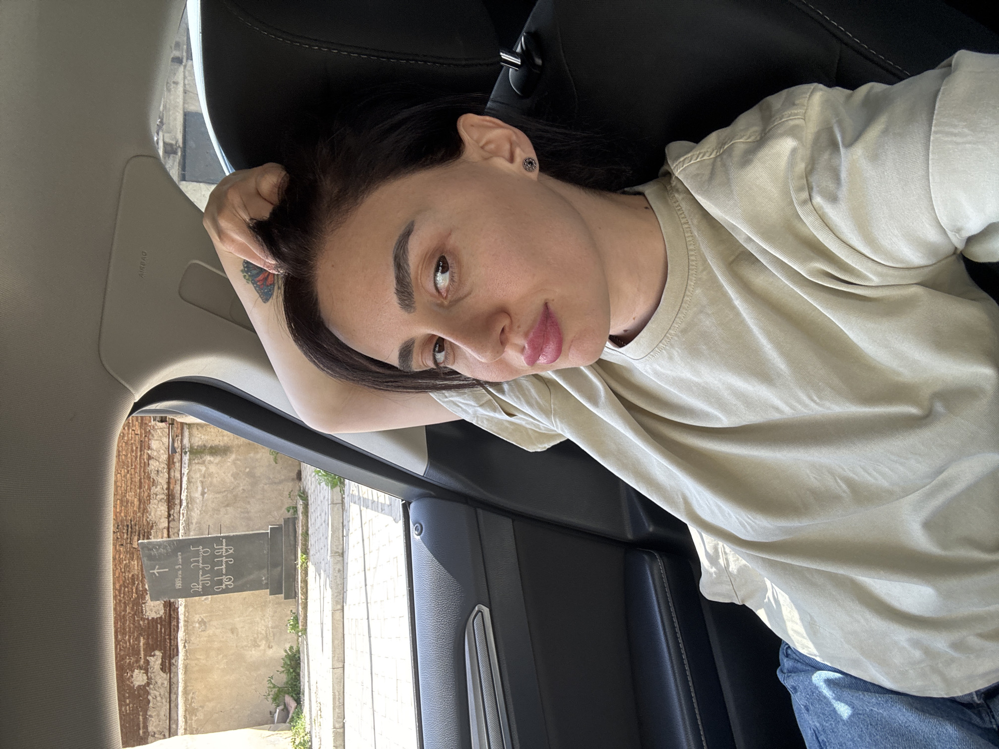 A woman with dark hair and a light-colored t-shirt is reclining in the passenger seat of a car. She is wearing blue jeans. The car appears to be stopped near a cemetery, as a tombstone is visible through the window. The tombstone has Georgian inscriptions, and the location appears to be Tbilisi, Georgia based on architectural details and the language on the tombstone.