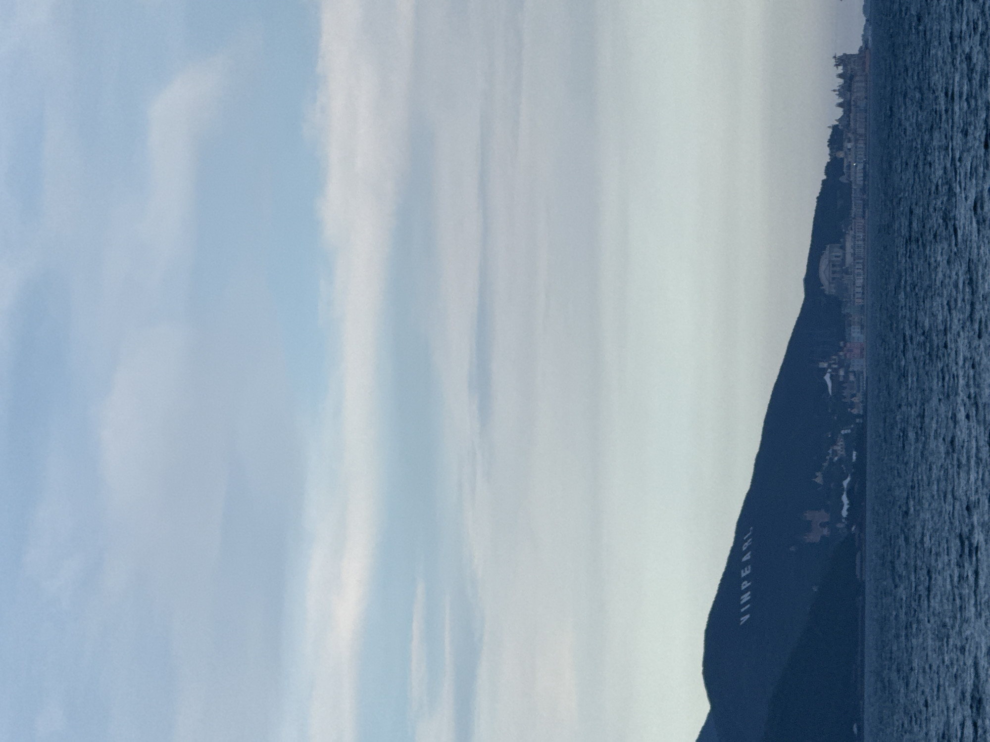 This media file depicts a coastal scene, likely in Nha Trang Ward, Vietnam. The image is oriented vertically, showing a body of water in the foreground and a dark, mountainous landmass on the right. The water is a deep blue, with visible ripples and texture, suggesting it is either the sea or a large lake.
The landmass is covered in dark vegetation and features a prominent inscription on its slope, which reads "VINPEARL". Above the landmass, a town or city is visible, with buildings clustered along the coastline and climbing up the hillside. A large, white domed building is particularly noticeable among the urban structures. The sky above is a pale blue with streaks of white clouds, indicating daylight conditions. The overall mood of the image is serene and perhaps a bit hazy. The time of day appears to be late morning or early afternoon, given the lighting. The location cues strongly suggest a tourist resort area given the prominent branding on the landscape.