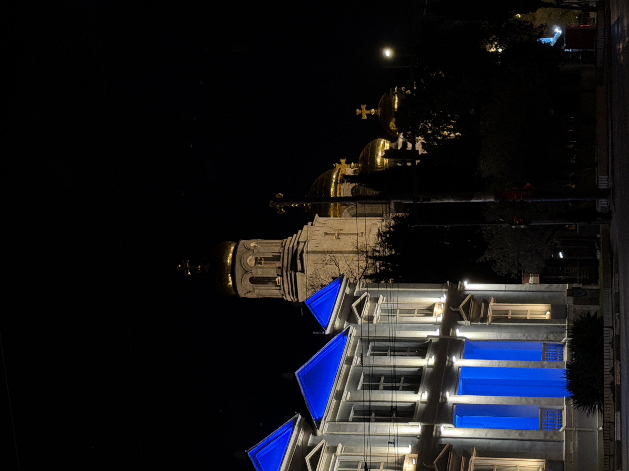The image shows the Cathedral of the Assumption of the Virgin in Varna, Bulgaria, at night. The cathedral's golden domes are illuminated and visible against the dark sky. The lower part of the image displays a building with blue-lit rooftop architectural details. Trees and a street can be seen on the right side. A street light is shining.