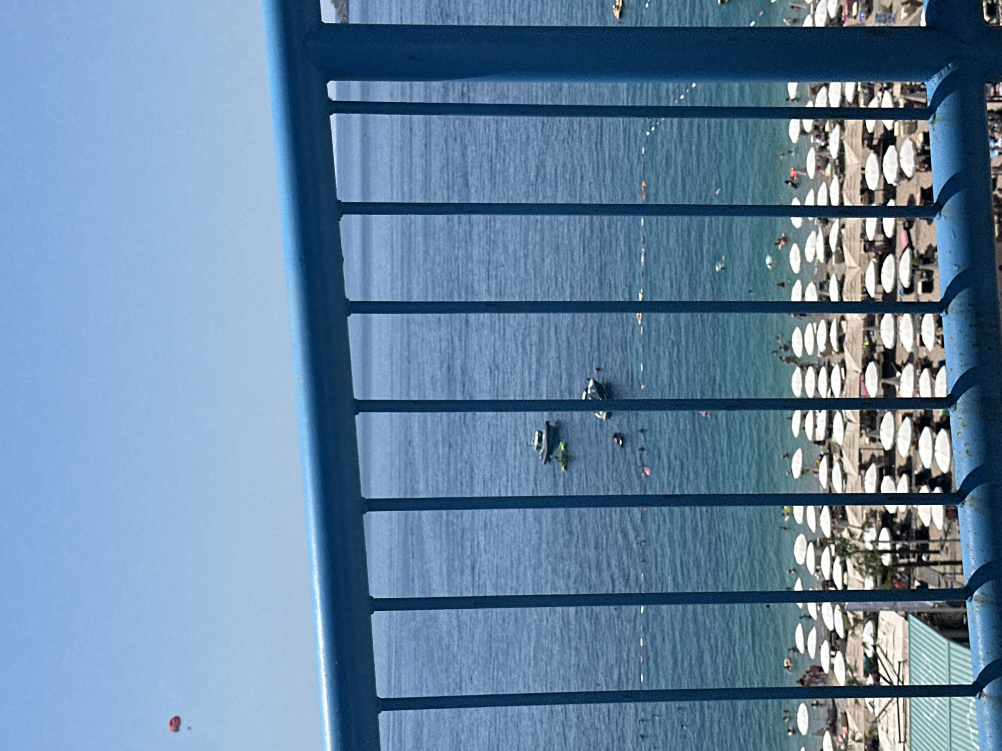 In Kamenovo, Montenegro, this image captures a vibrant beach scene on a clear, sunny day. A bright blue metal railing forms the foreground, with vertical bars obscuring a portion of the view. Beyond the railing, the azure Adriatic Sea stretches to the horizon. Several boats dot the water, including a small, dark-hulled vessel and a lighter-colored boat. Numerous beach umbrellas, predominantly white, are spread across the sandy shore, indicating a popular tourist destination. The beach is populated by many people, some swimming or wading in the shallow water, while others are relaxing under the umbrellas or strolling along the sand. In the upper left corner of the frame, a red parasail is visible against the light blue sky, suggesting recreational activities. The overall atmosphere is one of a lively summer day at the beach.