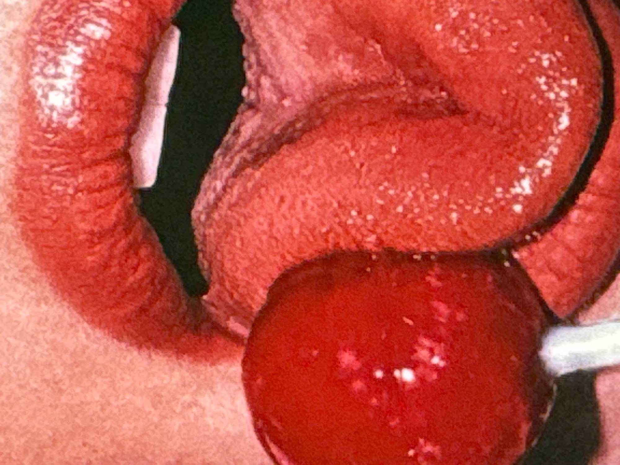 The image is a close-up shot of a person's mouth and tongue interacting with a red lollipop. The lips are painted red and are parted, revealing teeth. The tongue, also red and textured, is extended and touching the lollipop. The lollipop itself is round and a bright red color, with a white stick. The background is out of focus and appears to be skin texture. There is no indication of the city of Kuta Selatan, Indonesia, or any other environmental details.