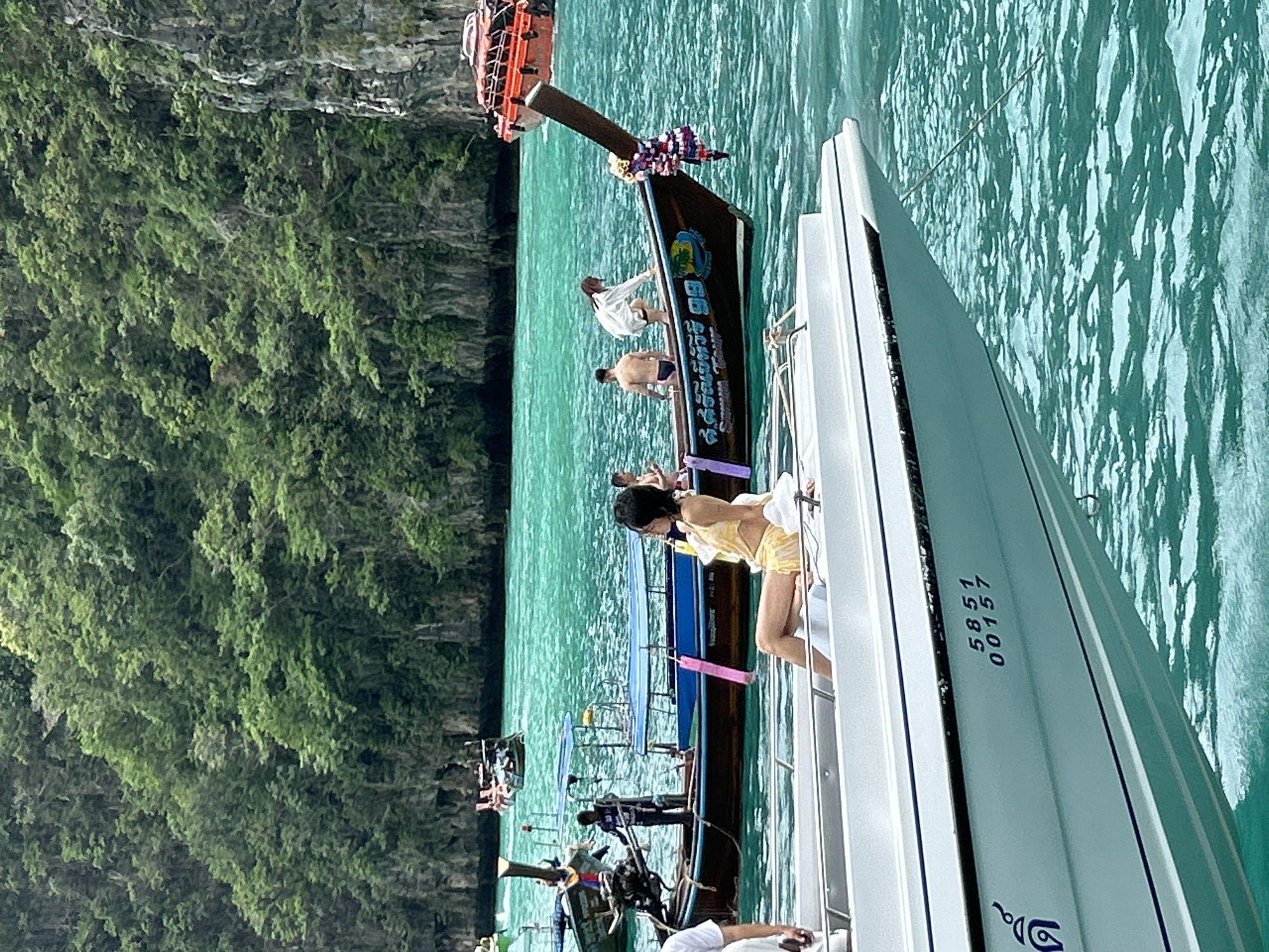 In Ao Nang, Thailand, a fleet of long-tail boats and speedboats navigate the turquoise waters near limestone karsts covered in lush greenery. A woman in a yellow bikini sits on the edge of a white speedboat in the foreground, facing away from the camera. Behind her, a traditional long-tail boat carries several passengers, including a man in blue shorts and a woman in a white top and black shorts who is leaning over the side. Another long-tail boat, slightly further back, has a man standing at the stern, steering. In the distance, more boats are visible, along with the imposing rock formations characteristic of the Thai coastline. The water is calm and clear, reflecting the vibrant green of the foliage.