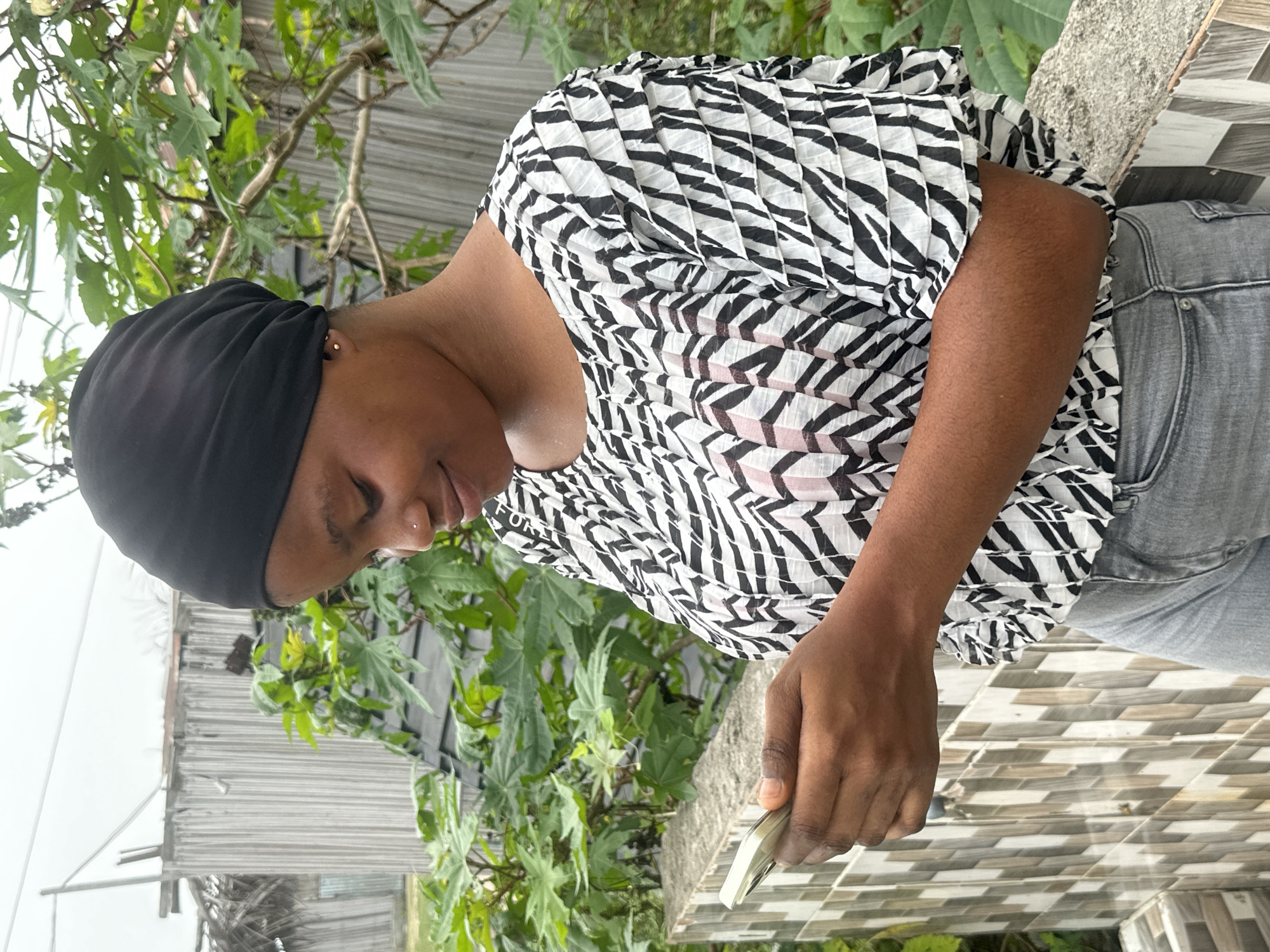 A young woman stands outdoors in Badagry, Nigeria, under an overcast sky. She is depicted with her head slightly tilted downwards, gazing towards a smartphone held in her right hand. She wears a black head covering, a black and white pleated top with a geometric, possibly zebra-like, pattern, and grey denim jeans. The partial word "FOR" is visible on her top.
Her expression is gentle, with a slight smile. She has a small gold stud earring in her right ear and a tiny silver stud nose piercing. The background features abundant green foliage, suggesting a garden or natural environment, and a building with corrugated metal siding. To her right, she appears to be leaning against or standing next to a tiled surface with a grey and white patterned design. The overall setting implies a casual, daytime outdoor scene.