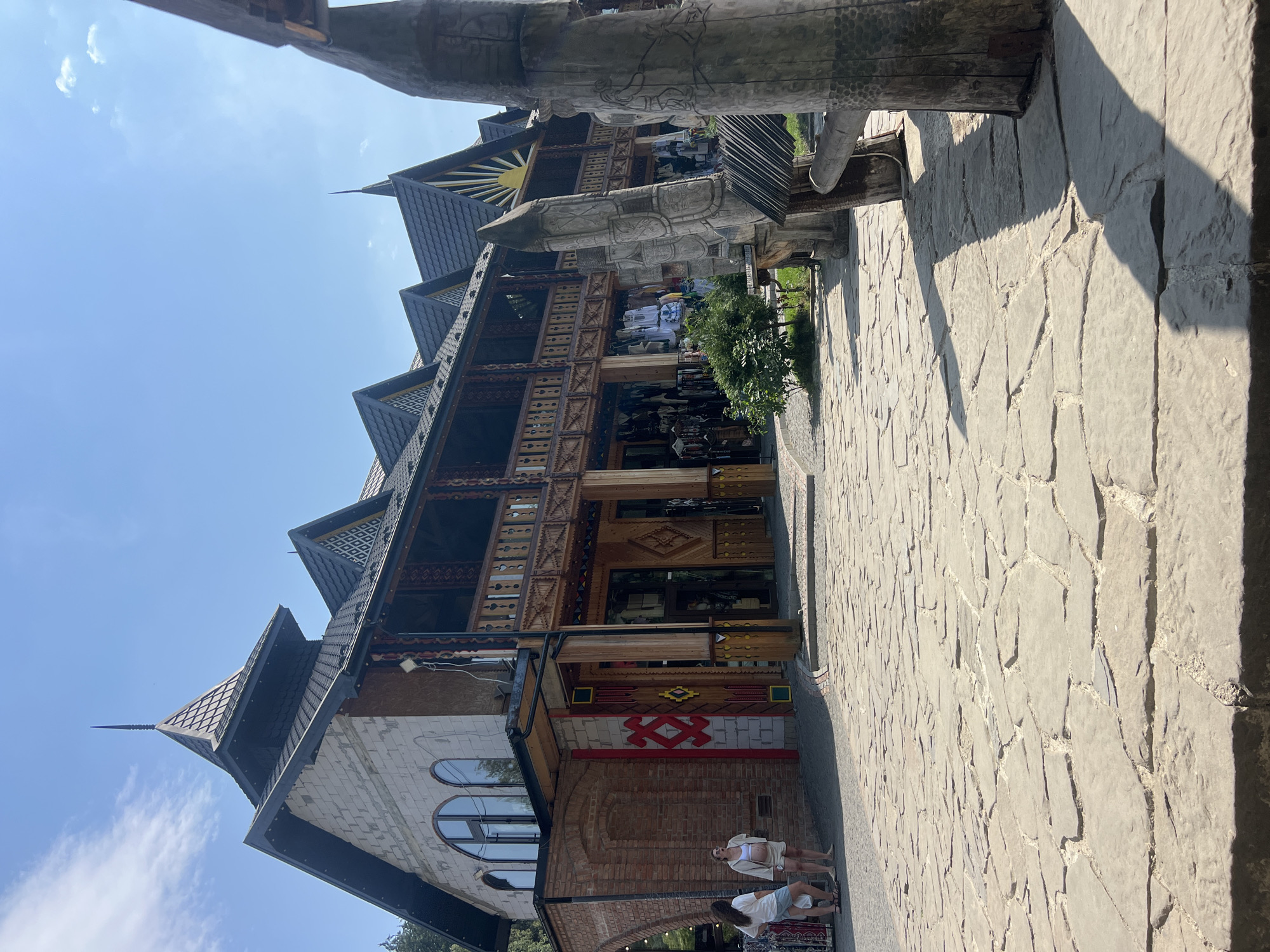 In Yaremche, Ukraine, a bright, sunny day unfolds in front of a multi-level building with a distinctive architectural style. The structure features a steeply pitched, dark-tiled roof with multiple gables and a central tower topped with a spire. The facade is a blend of stone, brick, and ornate wooden detailing, particularly along the second-story balcony. The building appears to be a marketplace or a series of shops, as evidenced by the displays of clothing and goods visible through open doorways and on racks outside.
The ground in front of the building is paved with large, irregularly shaped stones. To the right of the image, large, intricately carved wooden pillars stand, adding a rustic and possibly symbolic element to the scene.
Two women are standing near the entrance on the left side of the building. One is wearing a white t-shirt and shorts, and the other is wearing a light-colored jacket over a white top and shorts. They appear to be casually interacting or observing the surroundings. The sky above is a clear, bright blue with a few wispy clouds, indicating fair weather. The overall scene suggests a tourist destination with a focus on traditional or local crafts and architecture.