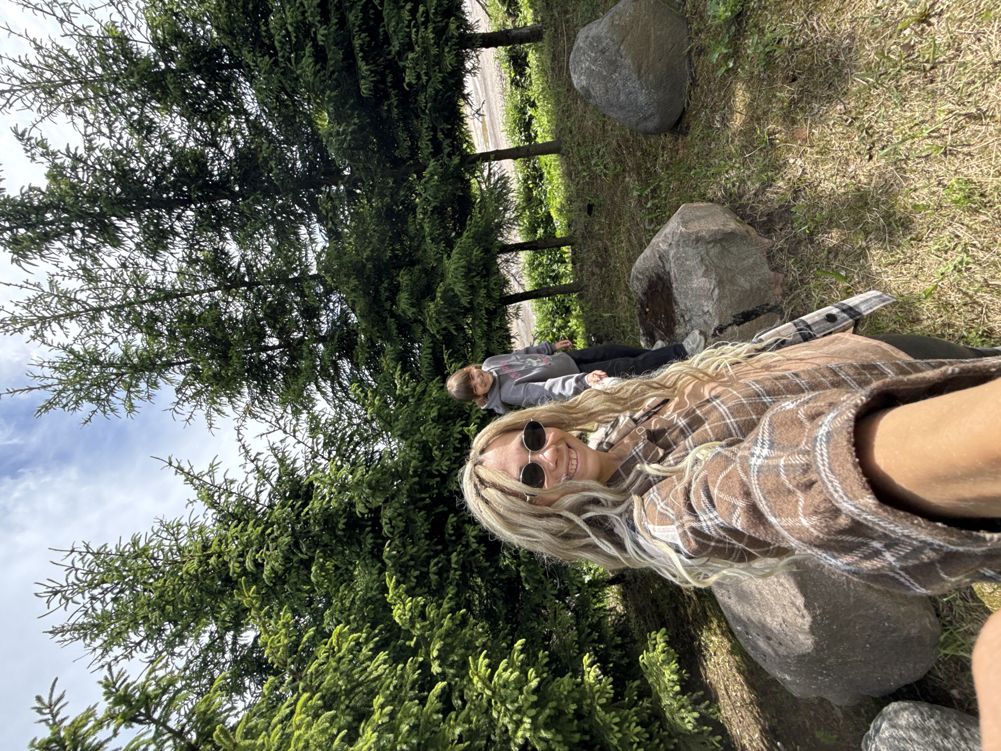 The scene depicts two people outdoors. A woman with long blonde hair wearing sunglasses and a brown plaid shirt is in the foreground, angled to suggest she is taking a selfie. Behind her, a child or young adult is lying down, facing the same direction, wearing a gray sweatshirt. The setting includes evergreen trees, a grassy area with scattered large gray rocks. The background features other trees and a hint of an asphalt surface suggesting a road or path. The location is likely Rusanovo, Russia, indicated by context.