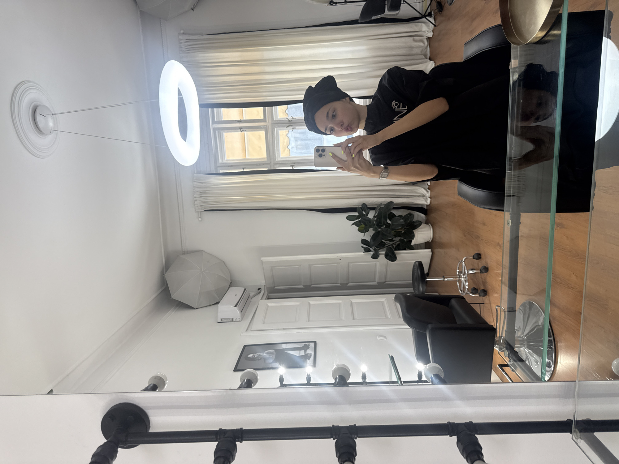 A woman wearing a black turban and black shirt is taking a selfie in what appears to be a room in the city of Unknown, Unknown. She is holding a smartphone in her hands. The room features a large window with white curtains, a circular light fixture suspended from the ceiling, a black salon chair, a glass table, a potted plant, and various other furnishings and lighting fixtures. Some of these elements are reflected in a mirror, creating a reversed view of the room.