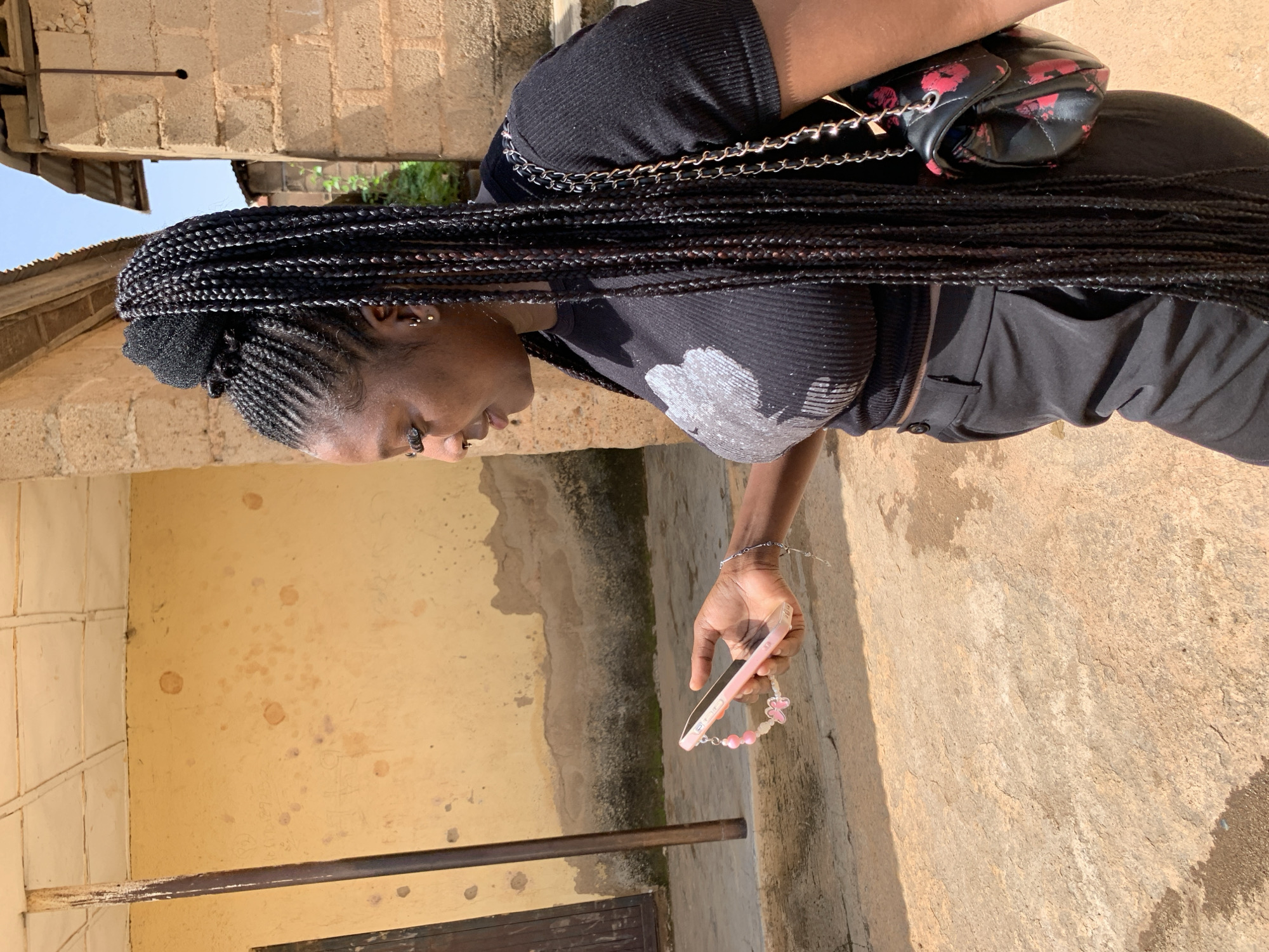 A woman stands outside in the daytime, holding a pink smartphone in her right hand. She is wearing a black crop top with a white graphic design and black pants. Her hair is styled in long, black braids that are gathered into a bun on top of her head. A black purse with a chain strap and a floral pattern is slung over her right shoulder. The background consists of a yellowish, textured wall with some smudges and marks, and a brick building with a corrugated metal roof. The ground is a rough, tan-colored surface, possibly dirt or concrete, with some dark wet patches. This scene is located in a residential or commercial area in Jos, Nigeria.