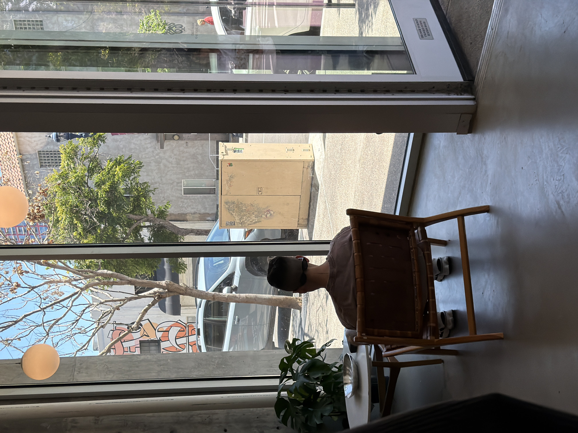 A person with short black hair, wearing a brown shirt, sits in a wicker chair next to a small white table with a potted plant. Two pairs of grey shoes are visible under the chair. The scene is viewed through a large window, revealing an outdoor cityscape. Buildings with graffiti and trees are visible across the street. A delivery box sits on the sidewalk. The location appears to be in an urban area of San Diego, California.