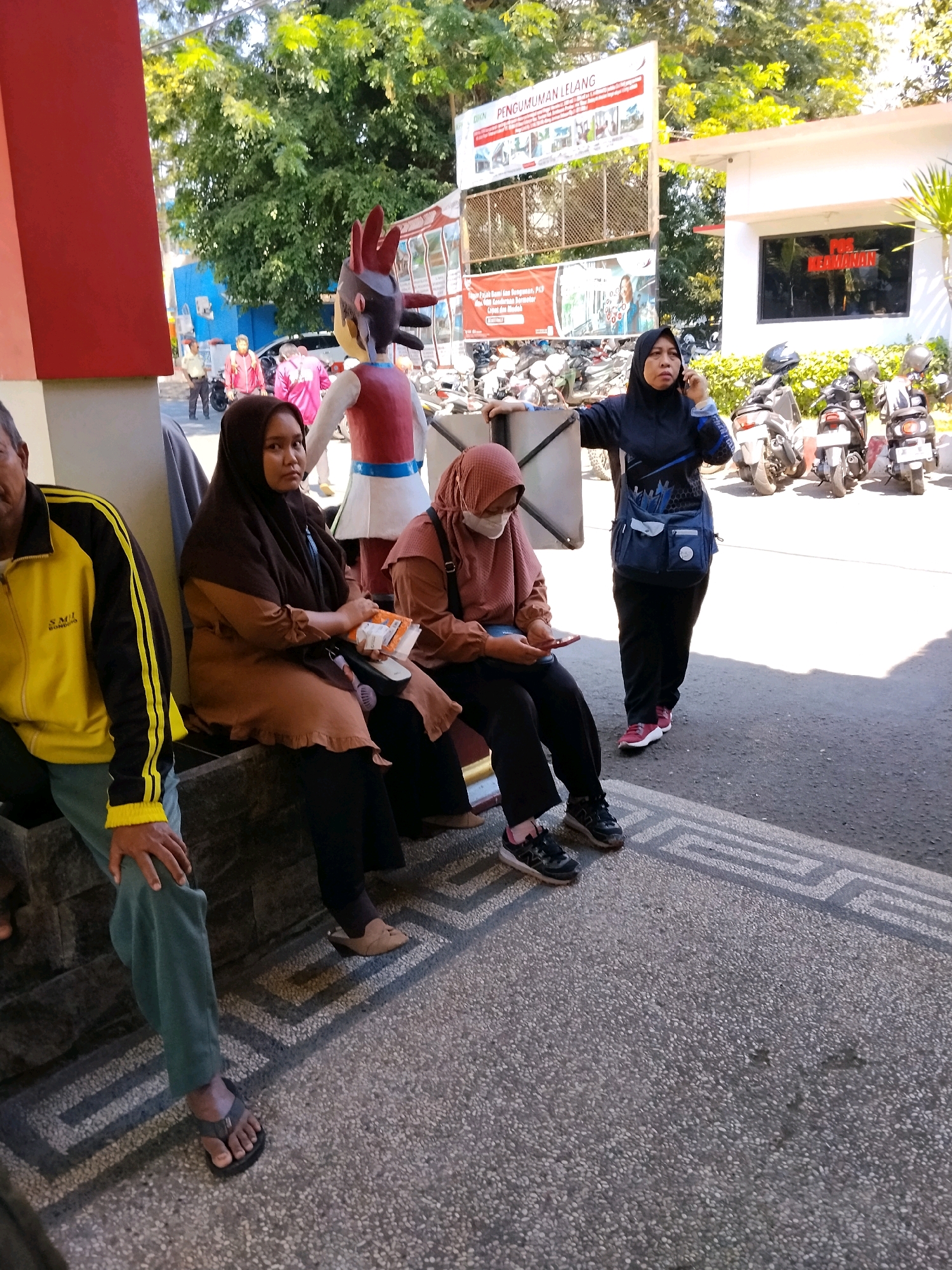 In front of a white building with glass windows displaying "POS KEDAMAIAN" signage, a woman in a dark blue hijab and jacket is speaking on her phone while holding a blue bag. To her left, two women are sitting on a stone bench. The woman on the right wears a brown hijab and a salmon-colored top, with a medical mask on her face and is looking at her phone. The woman to her left wears a brown hijab and a brown top and is holding some papers. To the far left, a person wearing a yellow and black jacket and teal pants is sitting with their hand on their leg. In the background, motorcycles are parked, and trees line the area. A statue of a cartoon character stands near the entrance of the building. The ground is paved with gravel and decorative tiles. The scene appears to be an outdoor waiting area, possibly at a government or public service building, given the signage and the presence of people waiting. The location is identified as Bondowoso, Indonesia.