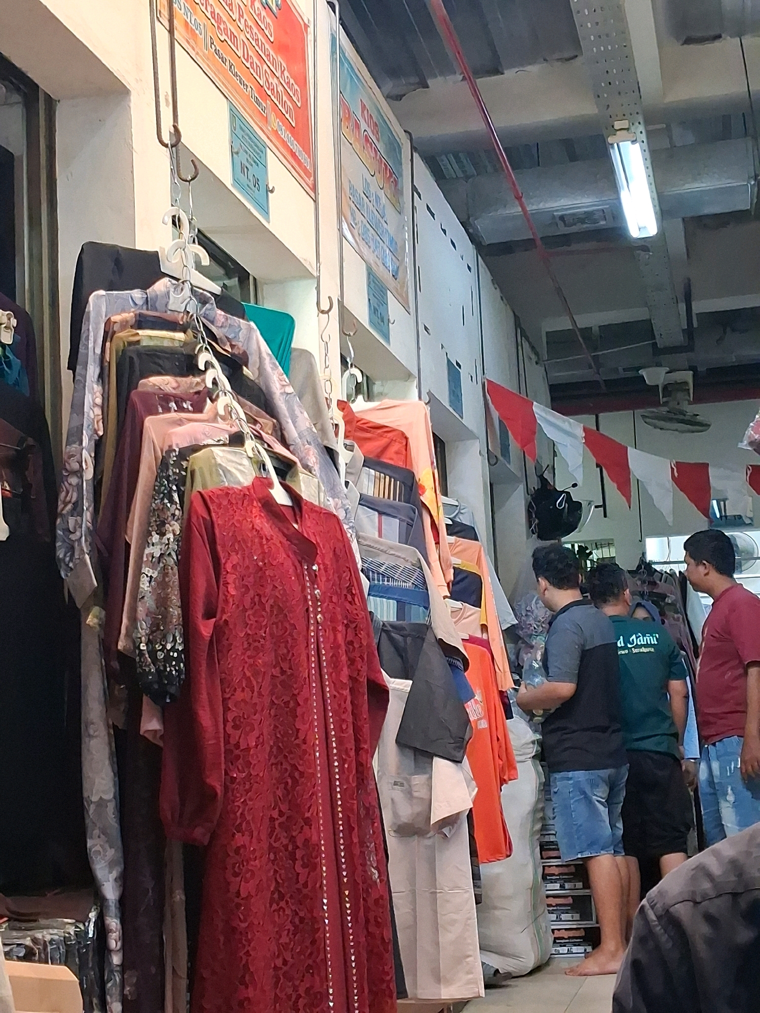 The image shows a view inside a marketplace in Surakarta, Indonesia, likely a clothing store. A rack of assorted women's clothing, predominantly blouses and dresses, hangs from metal hooks. A prominent red lace dress is displayed at the front. To the right, three men are visible, appearing to be customers or vendors. One man in a maroon shirt is looking towards the clothing, while another in a grey and green shirt is facing away, partially obscured. A third man in a maroon shirt stands to the far right. The environment appears to be an indoor market with fluorescent lighting and a ceiling with exposed ductwork and wiring. Red and white pennant banners are strung across the upper part of the scene, suggesting a festive occasion. Stacks of folded clothing or fabric are visible in the background. The overall impression is a busy retail environment typical of a local Indonesian market.