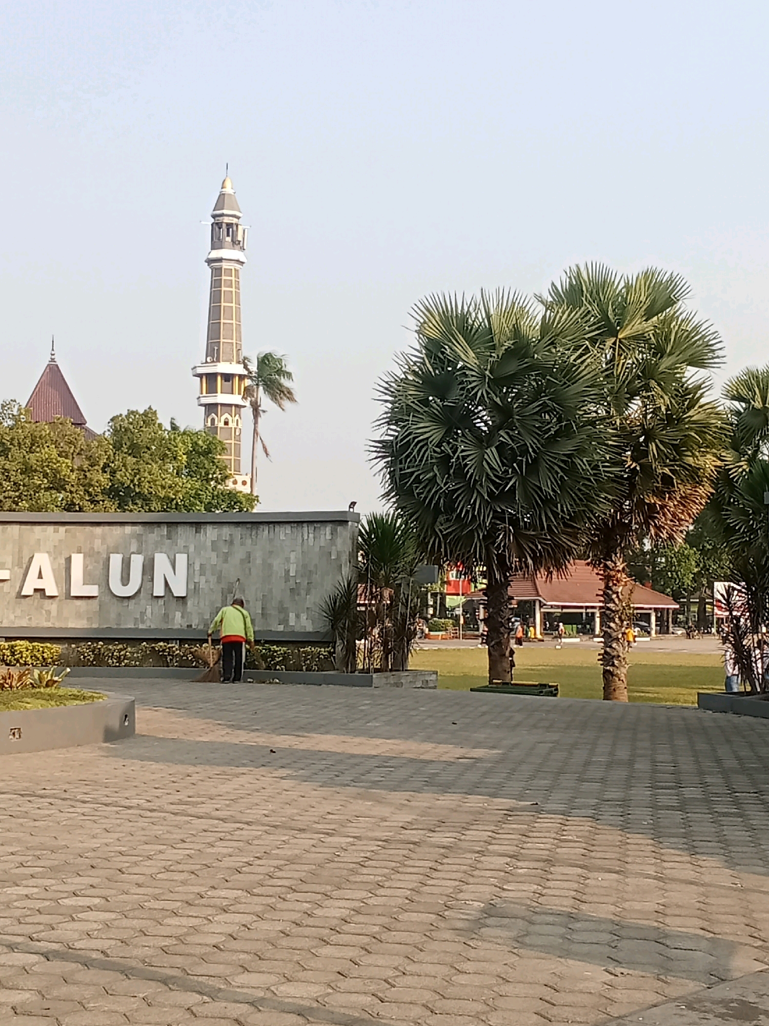 The image depicts an outdoor scene in Kaliwungu, Indonesia, featuring a man sweeping the pavement in front of a stone wall with large white lettering that reads "ALUN". In the background, a tall, multi-tiered minaret of a mosque is visible, along with lush green trees and palm trees. To the right, a brown-roofed building with open sides, resembling a pavilion or food stall area, is situated on a grassy field where several people are present. The foreground consists of a patterned gray brick pathway. The sky is clear and light. The time of day appears to be late afternoon, indicated by the soft lighting and shadows.