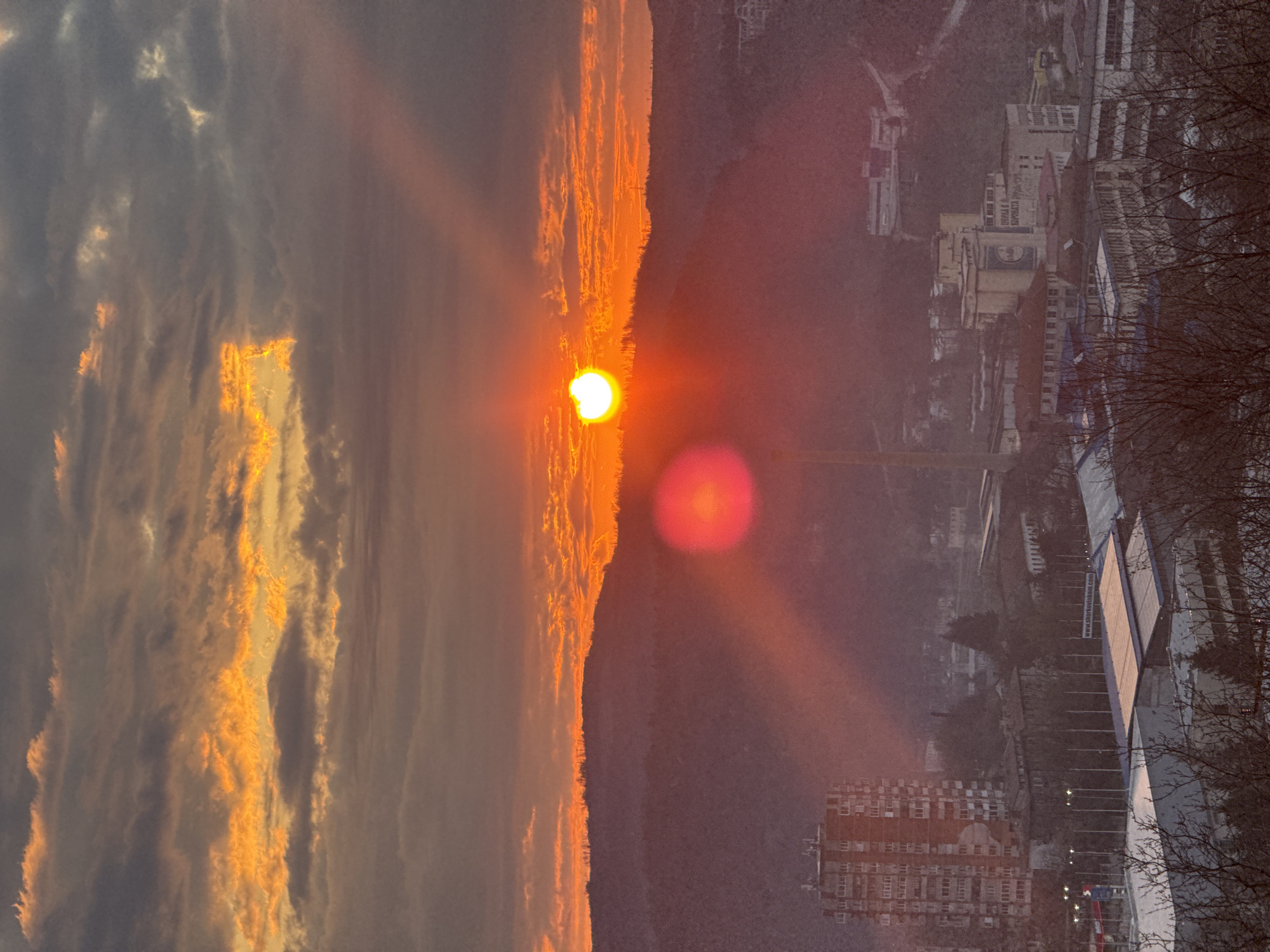 The image captures a sunset over Veliko Tarnovo, Bulgaria. The sun is positioned near the horizon line, emitting a bright orange glow that illuminates the surrounding clouds. To the right of the horizon, the cityscape is visible, including buildings, bare trees, and an elevated roadway with light poles. A tall construction crane is faintly discernible in the middle ground. The sky is filled with a mix of gray and orange clouds, with some clouds displaying golden hues from the setting sun. A reddish lens flare is noticeable near the sun.