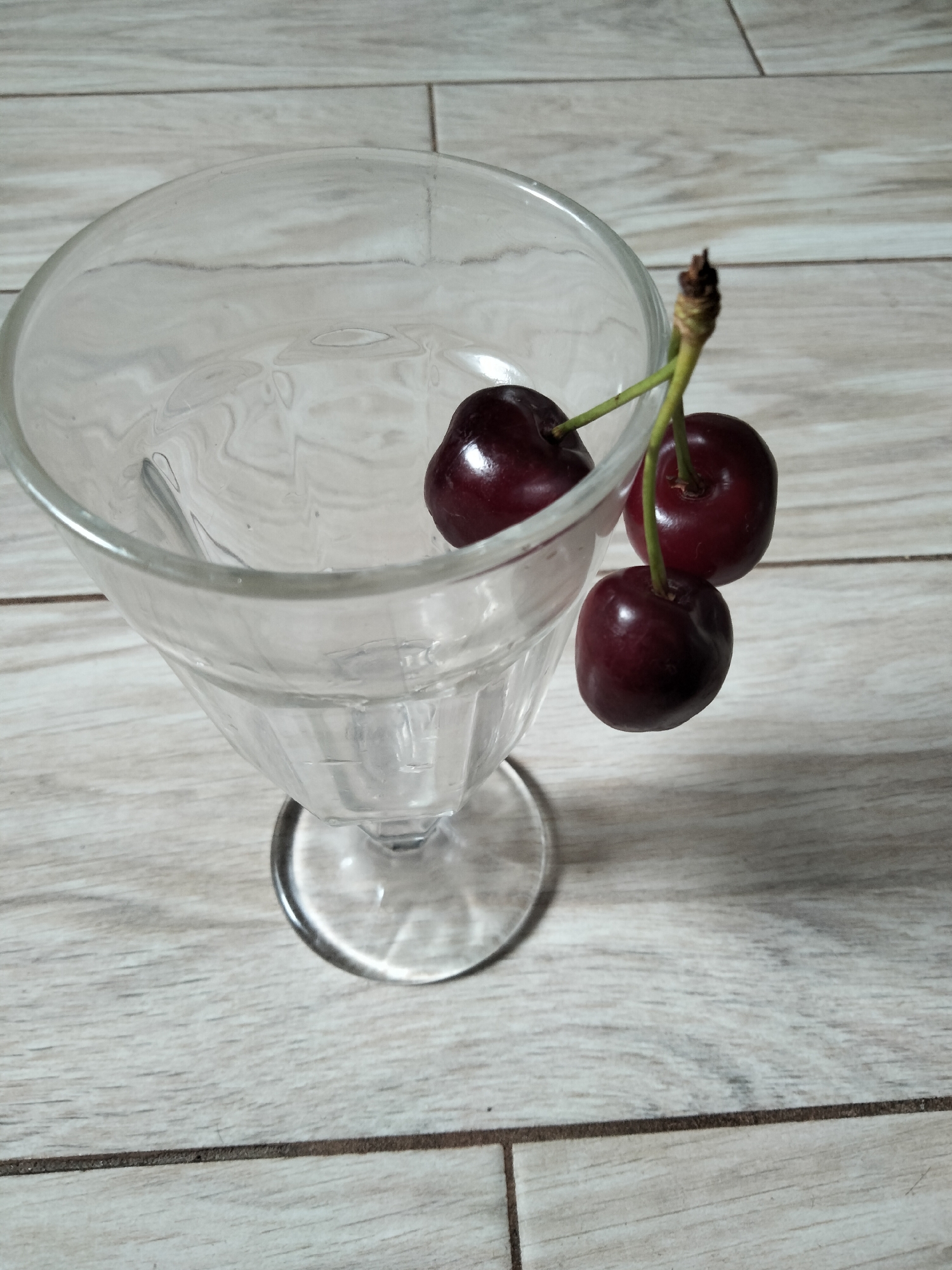 A close-up, eye-level shot shows a clear glass dessert dish with fluted sides and a stem, resting on a light-colored, wood-patterned surface. Three dark red cherries, still attached to their stems and one branch, are positioned to the right of the glass, with their stems partially inside the rim. The lighting is overhead and casts slight shadows. The image does not provide any visual cues to determine its location within the city of Kyiv, Ukraine.