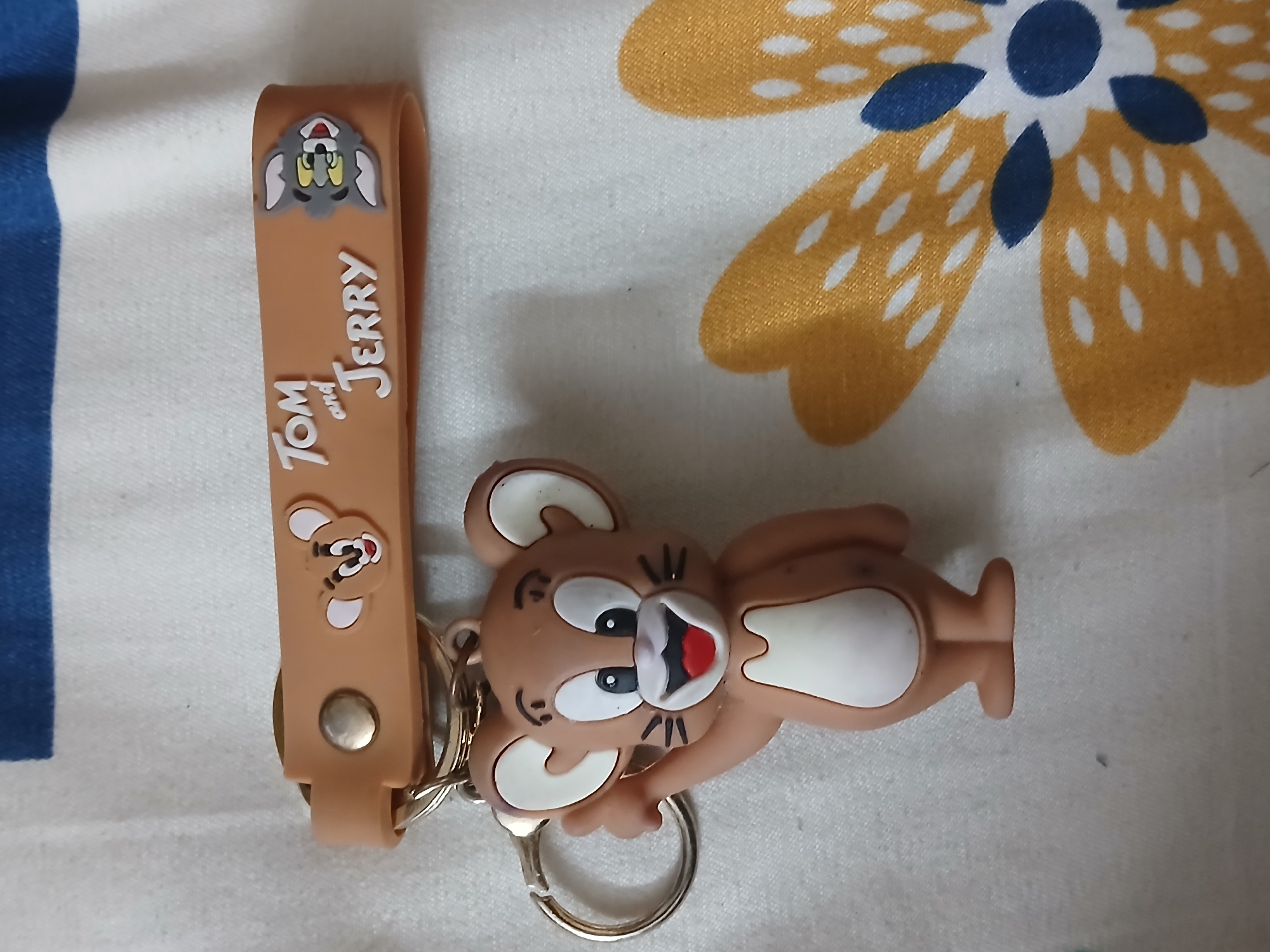 The image shows a Tom and Jerry keychain lying on a patterned surface. The keychain features a brown strap with "Tom and Jerry" written on it, along with cartoon images of Tom and Jerry. A Jerry figure is attached to the strap with a keyring. The background is a white cloth with a floral pattern in yellow and blue. The image was likely taken indoors. No people are visible. The location is unknown, but the request mentions the city of Bengaluru, India, which could be the place the image was taken.
