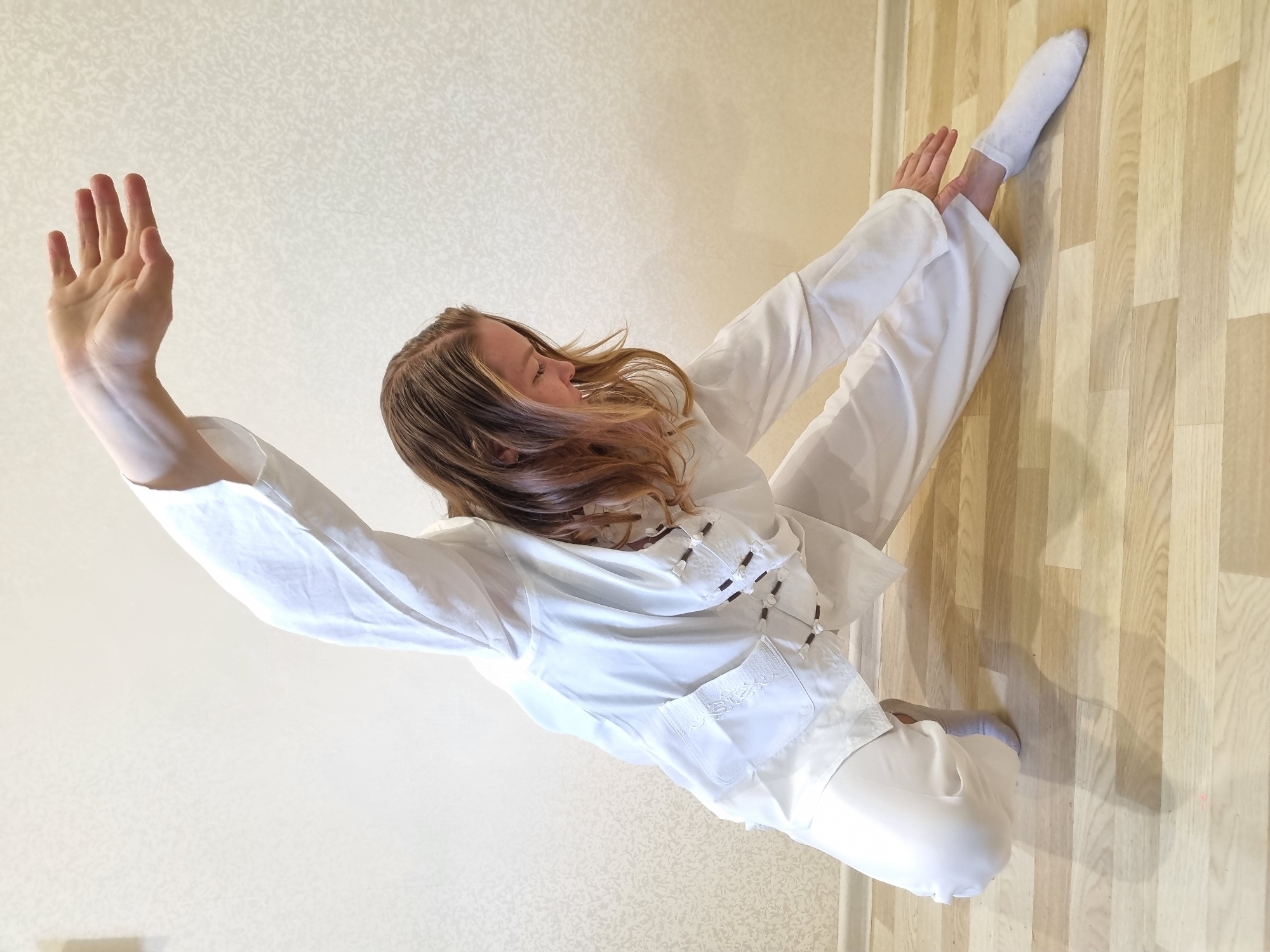 A woman is photographed from a high angle while practicing martial arts or a dance routine indoors. The location is likely in Chaikovsky, Russia given the prompt. She is wearing white martial arts clothing, including pants and a jacket with decorative closures. Her hair is brown, with lighter highlights. Her left arm is extended upward with her hand open, while her right arm is bent at the elbow, with her hand near her right leg. Her right leg is extended to the side, with a white sock on her foot. She is in a squatting position with her left leg bent. The floor is made of light wood planks. The wall behind her is textured and light-colored.