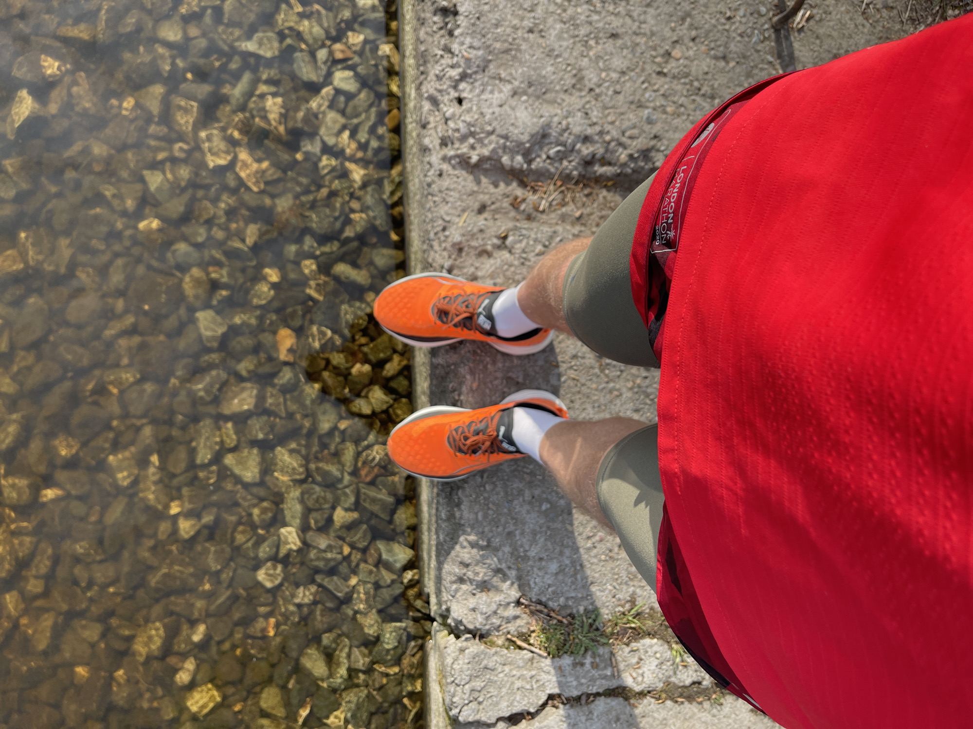 A high-angle shot captures a person standing on a concrete ledge next to a body of water, possibly near Izhevsk, Russia. The water is clear enough to reveal numerous small rocks on the bottom. The person is wearing bright orange running shoes with white socks, olive-green shorts, and a red running shirt or jacket. A small tag with "LONDON MARATHON 2020" is visible on the side of the shirt/jacket. The concrete ledge is gray and shows signs of wear and cracking. The lighting suggests it is a bright, sunny day.