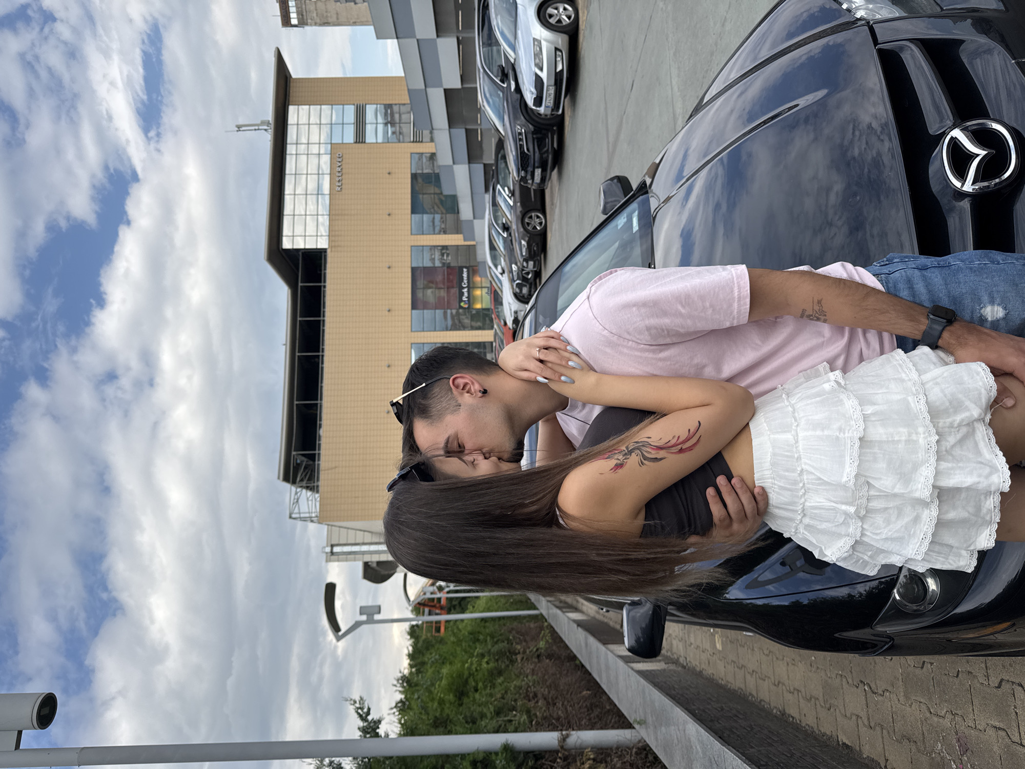 A young couple is depicted kissing in an outdoor parking area in front of a black Mazda vehicle. The female, with long dark hair, wears a dark sleeveless top and a white ruffled skirt, displaying a red and black phoenix tattoo on her right bicep and light blue nail polish. The male, wearing a pink t-shirt, blue ripped jeans, and a black watch, has sunglasses pushed onto his head, a small tattoo on his left forearm, and a black hoop earring in his left ear. They are embracing, with the female's right arm around the male's back and the male's left arm around the female's waist.
In the background, a modern multi-story building with a light brown facade and large reflective glass windows is visible. The word "RESERVED" is prominently displayed on the building's exterior, and "Park Center" is also visible, identifying the location as the Park Center shopping mall in Sofia, Bulgaria. A silver Skoda Octavia with a Bulgarian license plate "CA 4390 TC" is parked among several other cars to the right of the couple. The ground is paved with interlocking pavers, and tall modern streetlights are visible. Green trees and bushes line the left side of the scene under a partially cloudy sky.