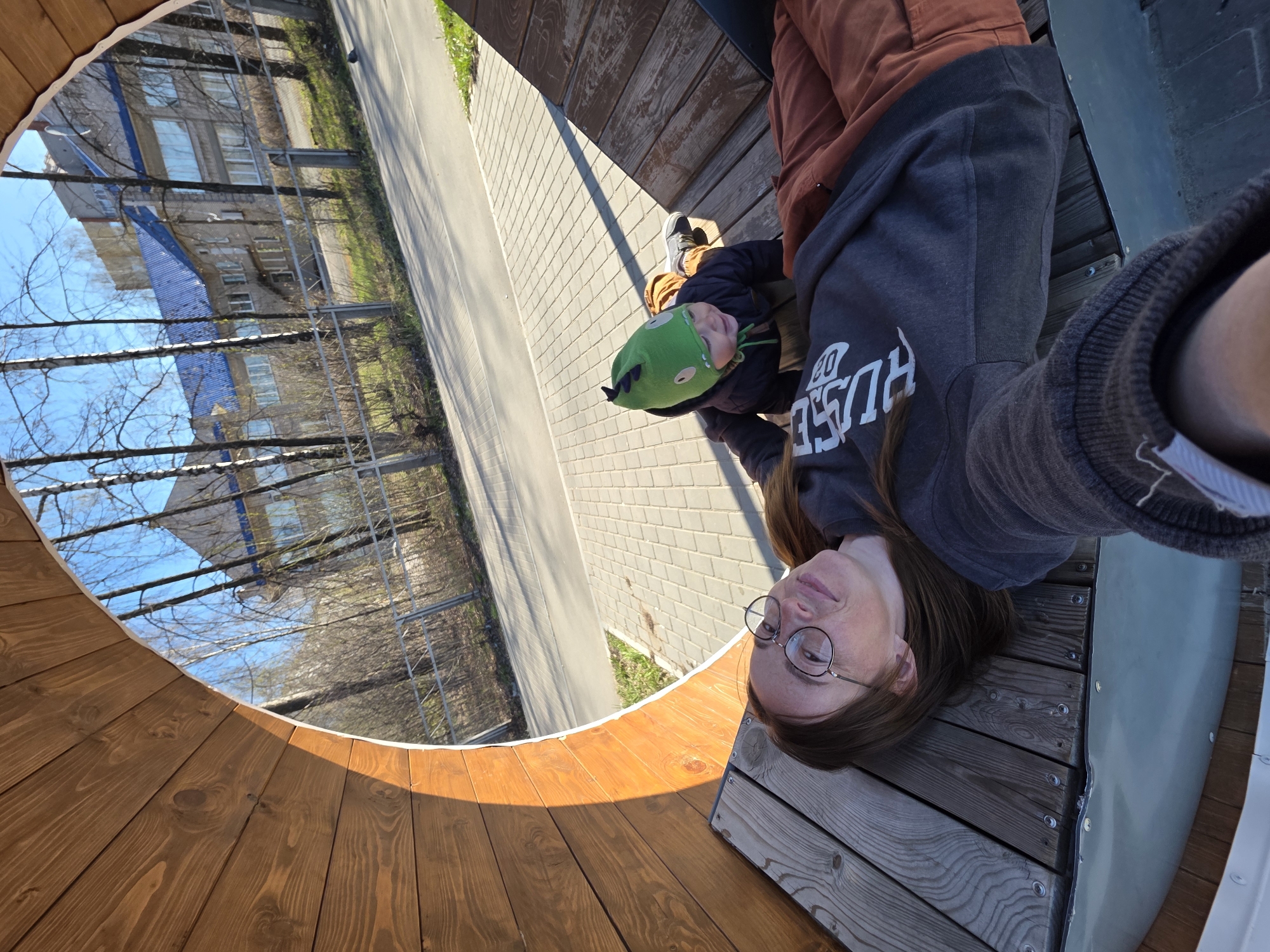A selfie of a woman in Chaikovsky, Russia, taken inside a wooden observatory structure, looking out at a street scene. The woman is lying on her side on a wooden bench within the structure, wearing glasses and a gray sweatshirt. A child wearing a green hat is partially visible. The observatory’s circular wooden frame provides a view of a paved road, buildings, and trees. The scene is sunlit.