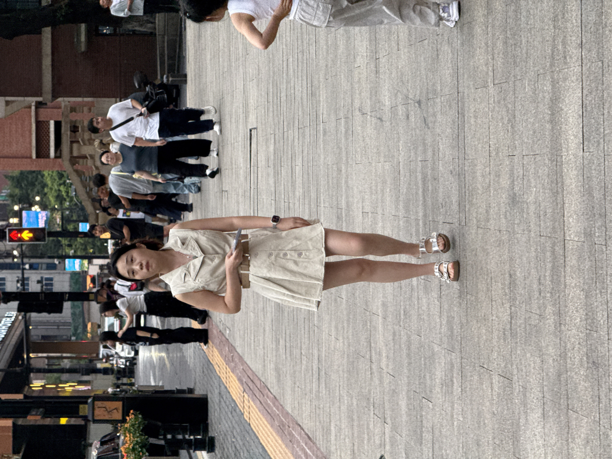 An outdoor medium shot captures a woman walking on a paved sidewalk in Yuzhong District, China. The woman, who is the primary subject, is wearing a light-colored sleeveless dress with a belted waist and silver sandals. She is looking forward and holding a rectangular object in her hands. In the background, other individuals are walking in various directions on the sidewalk, and to the left, there are buildings and parked vehicles. The environment appears to be a commercial or urban area, with a traffic light visible in the distance. The lighting suggests it is daytime.