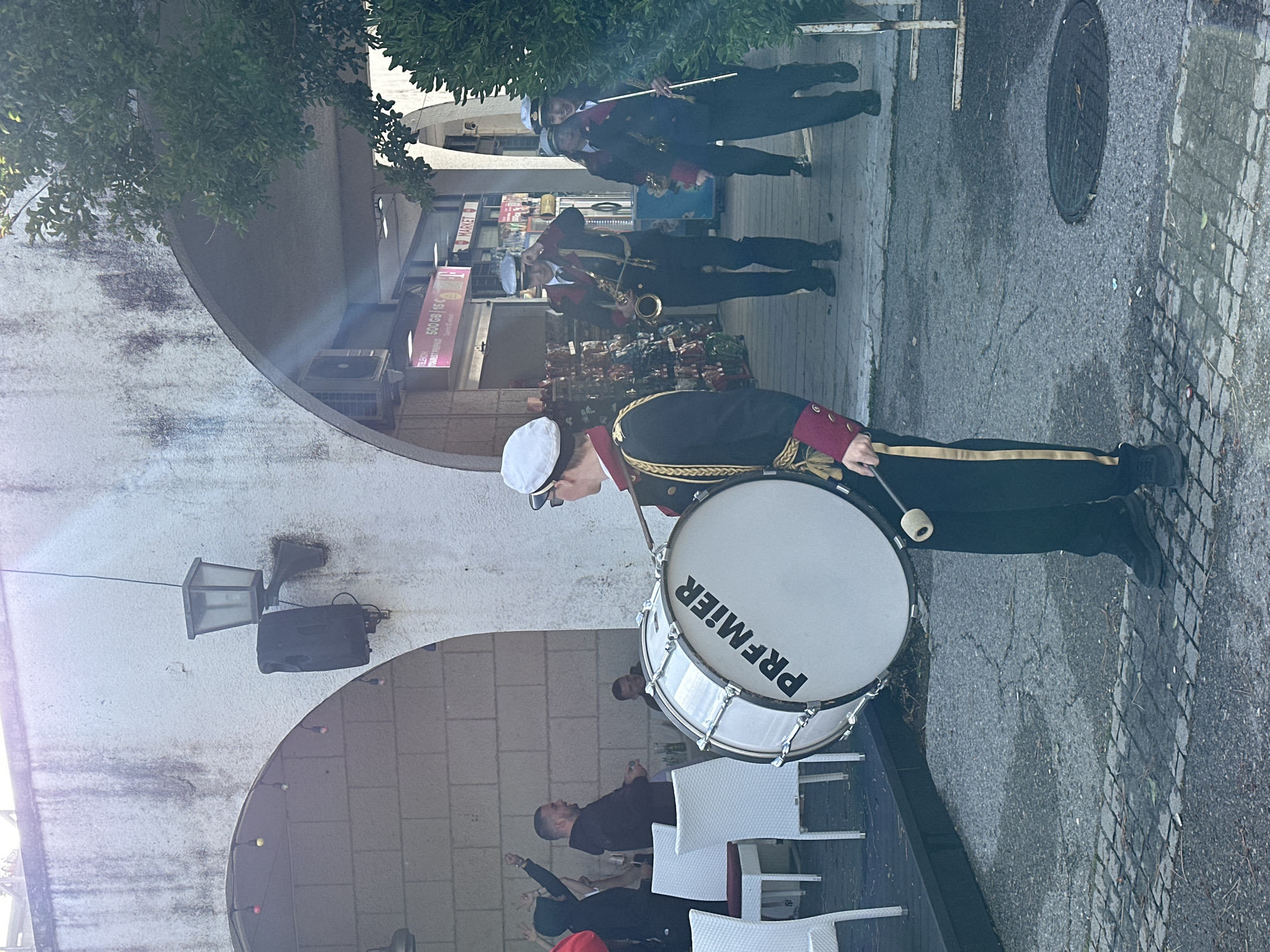 A street scene in Budva, Montenegro depicts a marching band. A drummer in a formal uniform stands at the forefront, holding a beater and a Premier drum. Behind him and to the right, a saxophone player and two other musicians are visible. The band appears to be marching past an outdoor restaurant area, evidenced by white chairs and tables. A stone building with arched openings and a light fixture serves as a backdrop. A manhole cover is visible on the street.