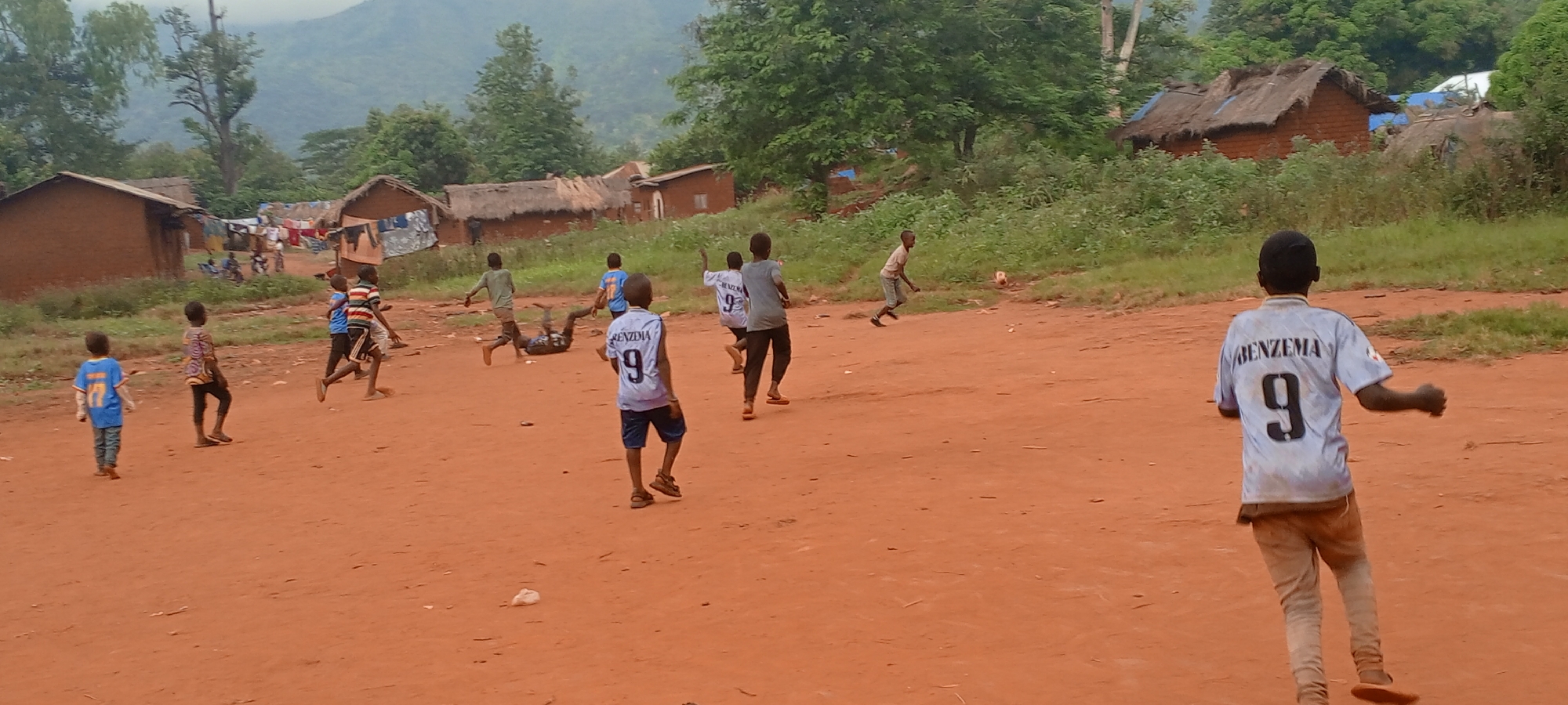 In Ngalula, Democratic Republic of the Congo, a group of approximately ten boys plays soccer on a dirt field. The boys, wearing a variety of casual clothing, including soccer jerseys, are in motion, running and kicking a soccer ball. In the background, simple brick dwellings with thatched roofs and clotheslines are visible, indicating a rural or impoverished setting. The landscape features rolling hills covered with lush greenery, suggesting a tropical climate.