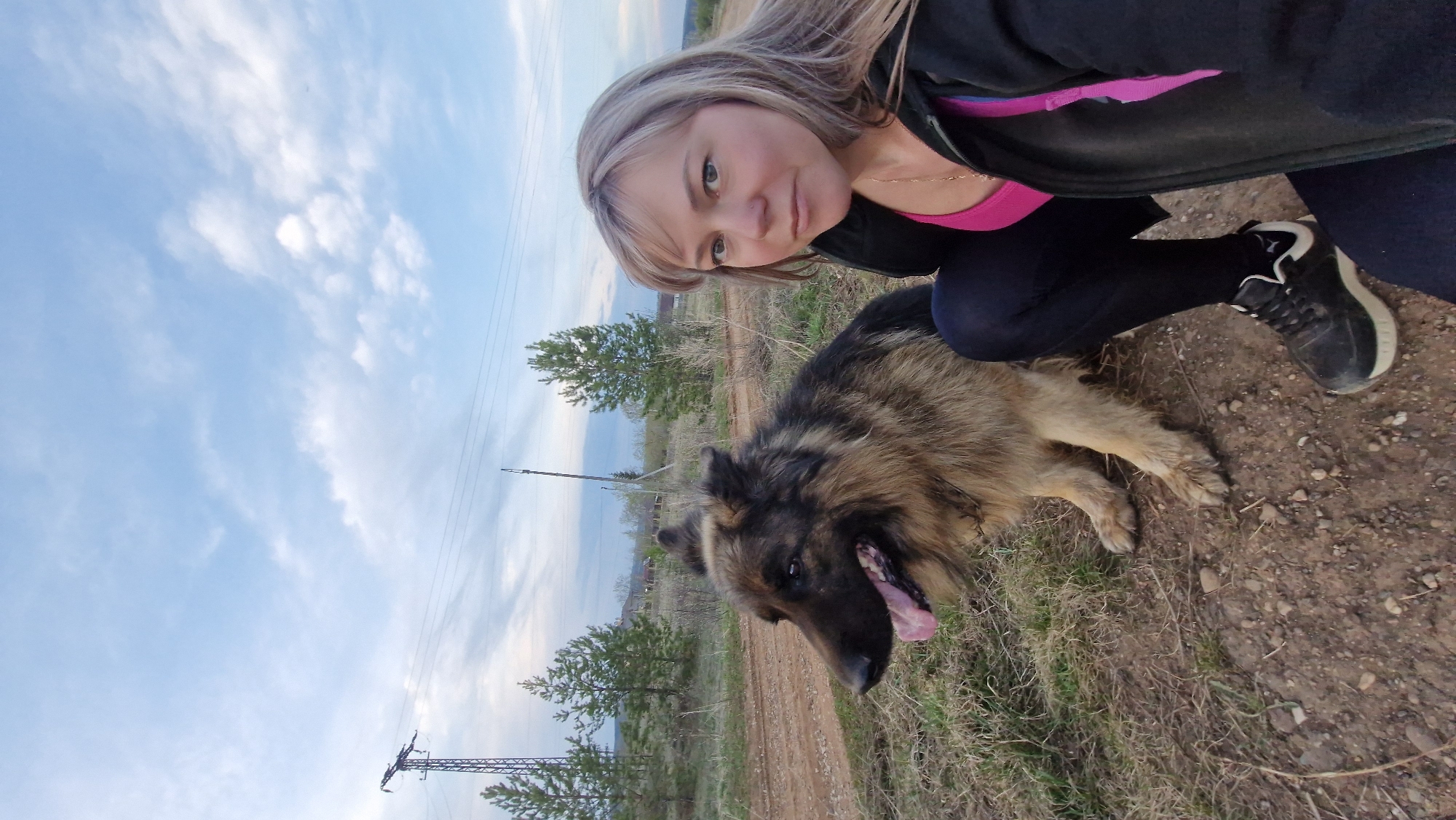 A woman and a large dog are in a field on the outskirts of Otchor, Russia. The woman, with blond hair and wearing a black jacket over a pink top and black leggings and shoes, is kneeling beside a German Shepherd-like dog with its tongue hanging out. The dog is lying on the ground, which is covered in dry grass. Overhead power lines are visible in the background. The sky is blue with clouds.