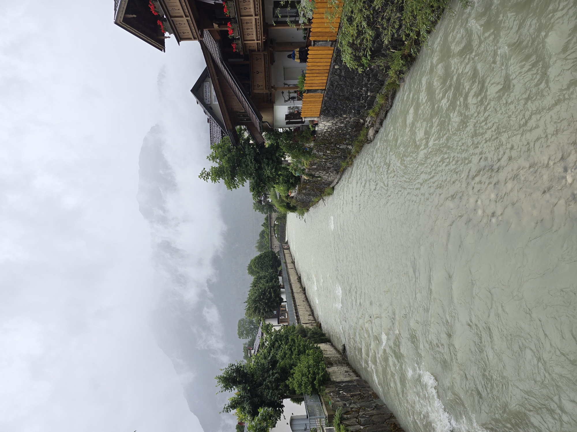 The image shows a view of the Loisach river in Garmisch-Partenkirchen, Germany, on a cloudy day. The river flows through the town, with buildings and trees lining its banks. In the foreground, the river is a murky, greenish-brown color, with visible ripples and currents. The left bank of the river has a stone wall with a metal railing, and beyond that, a white building with gray doors and trees. On the right bank, there are traditional Bavarian-style houses with wooden balconies and red flowers. A yellow fence separates the houses from the river. In the background, mountains are visible, partially obscured by clouds. A stone bridge can be seen in the distance. No people are clearly visible in the image, although a person in a blue and yellow jacket is partially visible on the right bank, near the yellow fence.