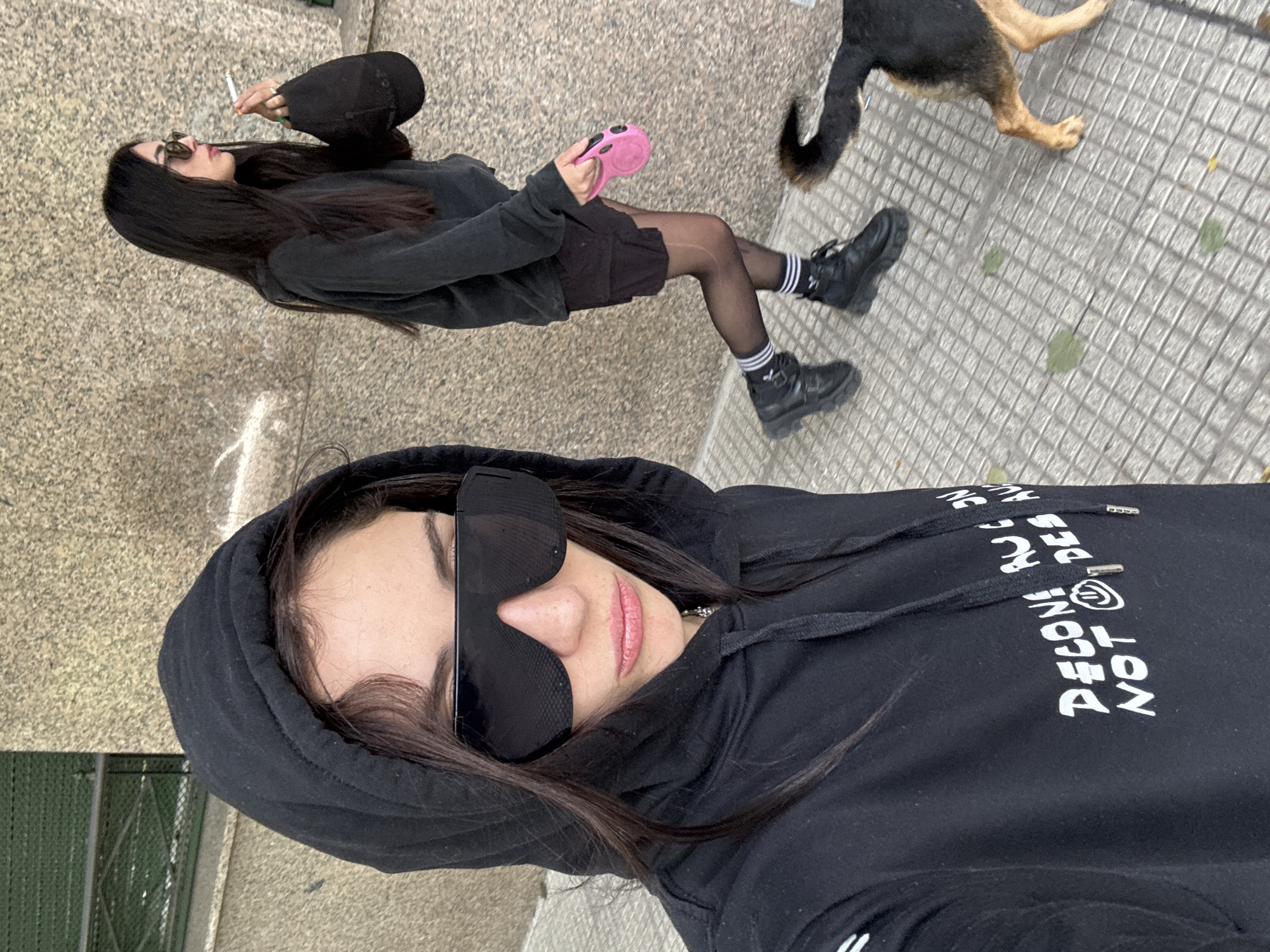The image appears to be a selfie taken outdoors on a city street in Buenos Aires, Argentina.
Two women are visible. One is in the foreground, wearing a black hooded sweatshirt with white lettering, and large sunglasses that cover much of her face. She is looking directly at the camera. The second woman is in the background, at a distance. She is dressed in black shorts, a black sweater, black tights, and black boots, and is wearing a black baseball cap. She is smoking a cigarette. A pink dog leash is in her hand.
The corner of a large dog is visible, likely belonging to the second woman.
The ground is paved with square tiles. There is a green metal fence and a section of sidewalk and street visible.