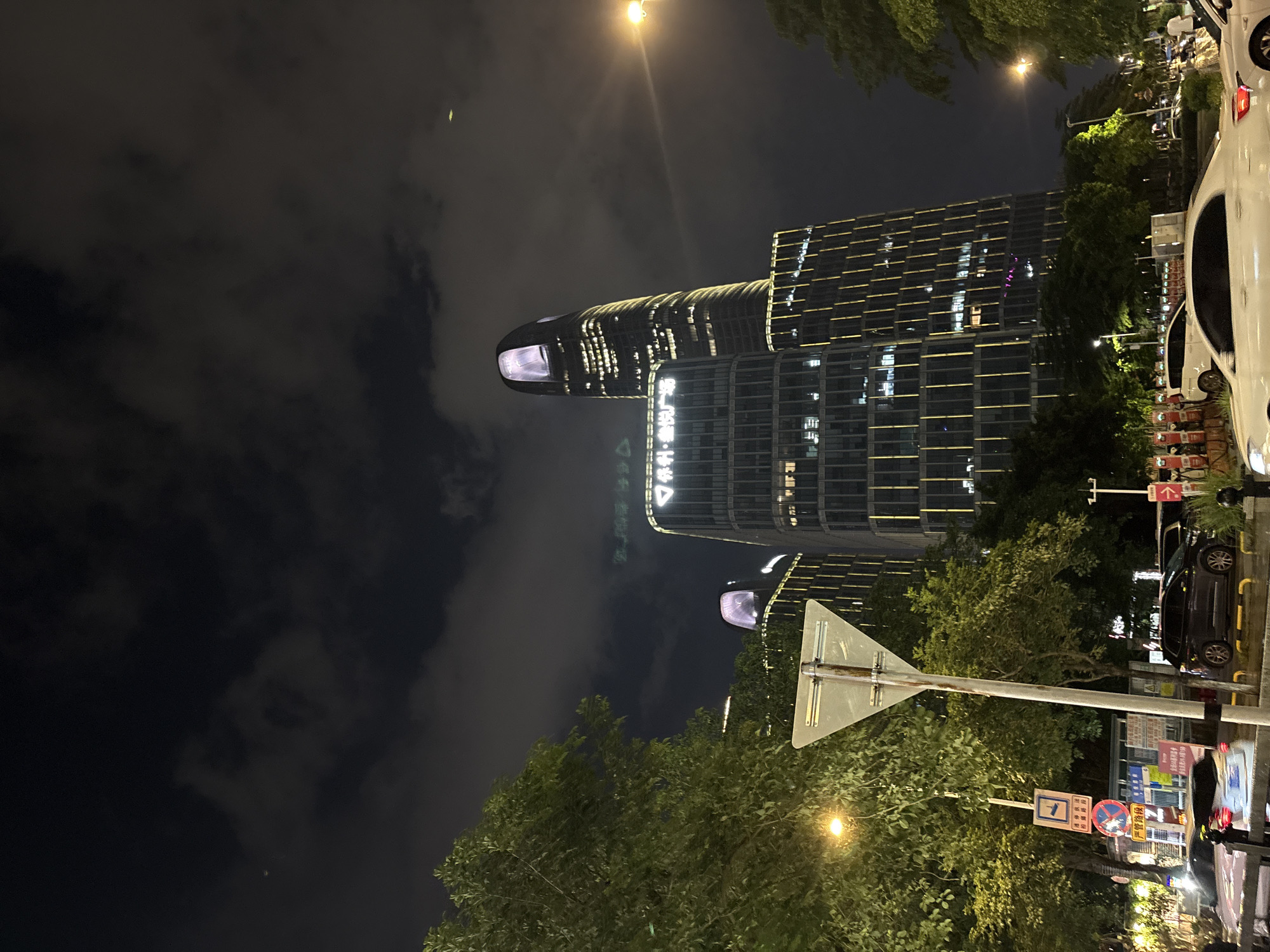 This is a night-time photograph taken in Haizhu District, China. The scene features modern architecture, including a distinctive building with a curved, illuminated facade. The building's lights create a warm glow against the dark sky, which is partially obscured by clouds. Trees with lush green foliage line the street, adding a natural element to the urban landscape.
In the foreground, a traffic sign with a reflective surface is prominently displayed. The sign has a triangular shape and is mounted on a pole. Several other signs with Chinese characters are visible in the background, some of which appear to be traffic or informational signs.
Several vehicles are present, including a white car on the right side of the image and a dark-colored car parked along the street. The presence of streetlights and the illuminated buildings indicate that it is nighttime. The overall mood of the image is one of a bustling urban environment at night, with a blend of natural and man-made elements.