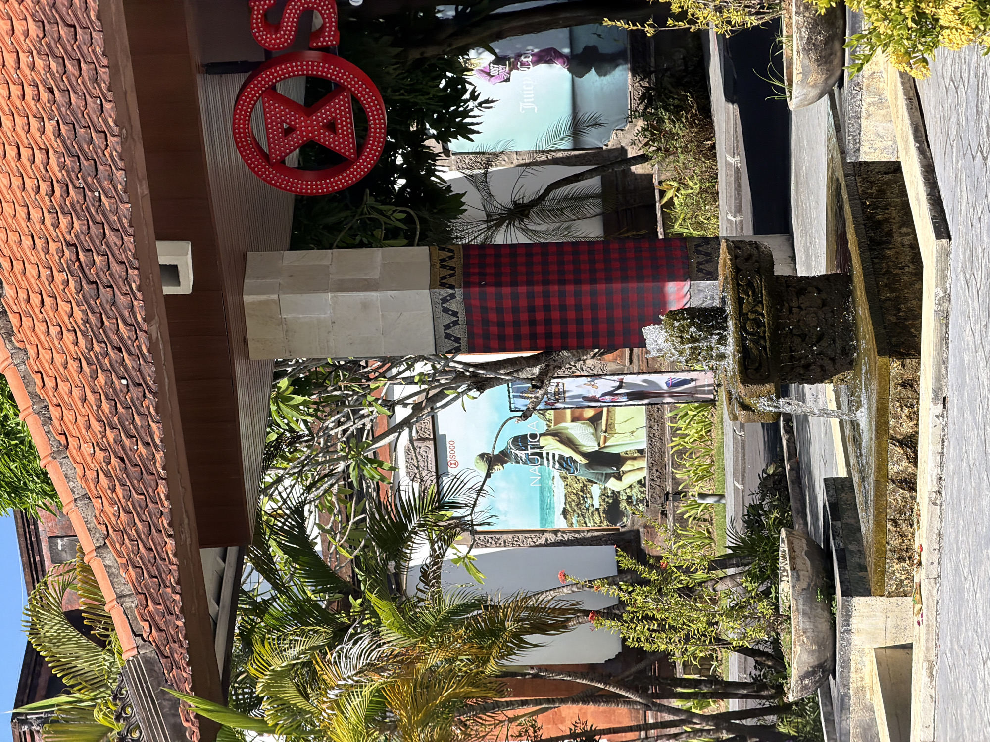 The image depicts an outdoor scene in Kuta Selatan, Indonesia, likely a commercial area. A tiled, terracotta roof extends over a paved walkway. In the foreground, a small stone fountain with water spouting is the central feature. To the left of the fountain, a large, weathered concrete planter holds a lush green plant. Beyond the planter, a building with a red tile roof is visible.
Dominating the mid-ground is a large column covered in red and black buffalo plaid fabric. Behind this, two large advertisements are displayed on white walls. The left advertisement features a male mannequin wearing a Nautica brand outfit, standing on a rocky shore with the ocean in the background. The right advertisement shows a female figure in a purple jumpsuit, holding a bag, with "Juicy Couture" text visible.
To the right of the plaid column and advertisements, trees with green foliage and a large red sign with white dots, forming the letters "X" and "OS," are present. The ground is paved with light-colored stone tiles, and a darker gray pavement is visible further back. Greenery, including palm trees, is interspersed throughout the scene, suggesting a tropical climate. The sky is clear and blue. The overall impression is of a commercial plaza or entrance area, possibly associated with a shopping center.