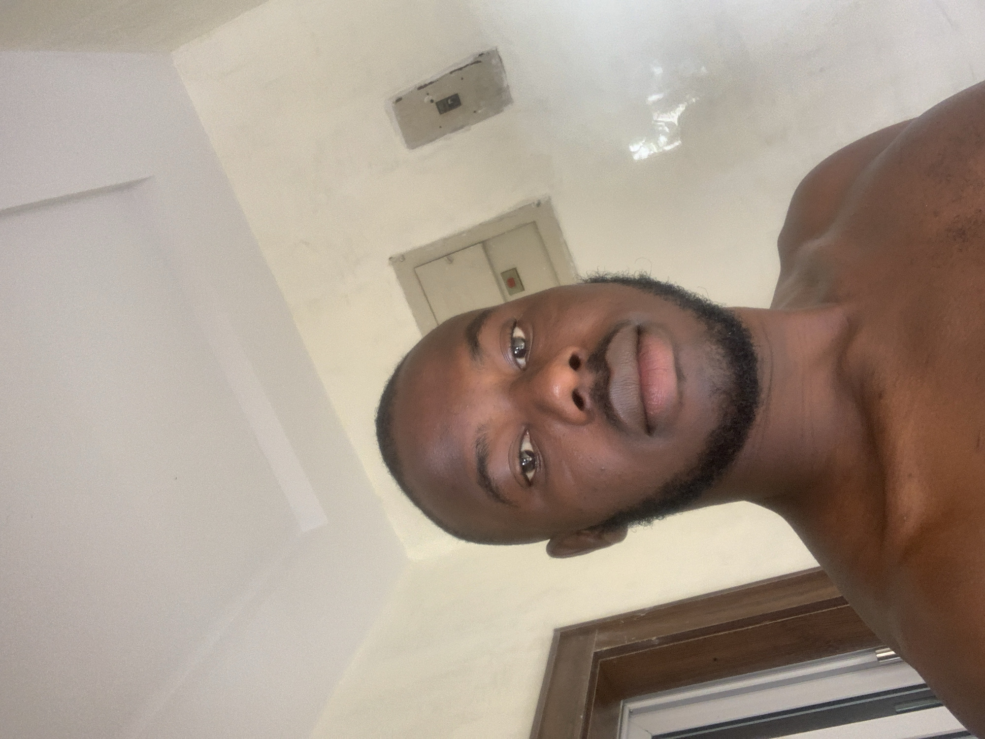 This close-up, upward-angled selfie features a dark-skinned man, likely in Jos, Nigeria, looking directly at the camera with a neutral expression. He has short dark hair, a neatly trimmed beard, and a mustache. Part of his bare shoulder and upper chest are visible on the right side of the frame.
The background consists primarily of a light-colored (off-white or cream) ceiling or upper wall, which exhibits some reflections, possibly indicating a slightly glossy surface or moisture. Two rectangular fixtures are visible on this surface: an upper, recessed electrical box or light fixture mount, and a lower panel resembling an electrical or access panel with a small red switch or indicator light. In the lower right corner, a dark brown wooden or metal frame, likely from a window or door, is partially visible, showing a pane of glass.
The lighting is bright and even. The image has a slightly grainy quality. The man is taking a selfie, capturing a personal perspective of himself and his immediate indoor surroundings. No visible text is present.