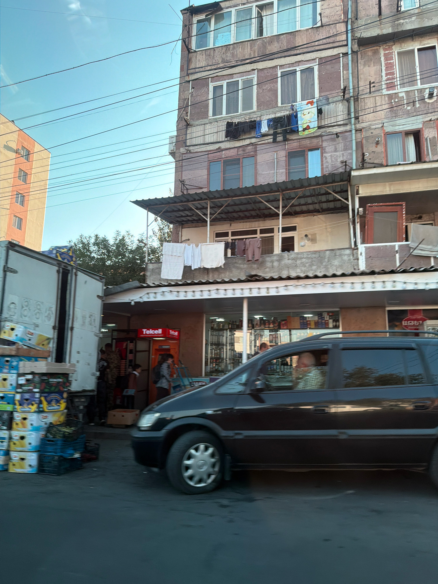 The image shows a street scene in Yerevan, Armenia, likely in a residential area. On the right, a multi-story apartment building with a pinkish-brown facade has clothes hanging from a line outside one of the windows on the second floor. Below the building, a storefront with a green awning is visible, with a "Telcell" ATM machine positioned outside. To the left of the building and storefront, a large white truck is parked, with its rear doors open and stacked boxes of produce, some appearing to be fruit, in front of it. A dark-colored minivan is driving past the scene on the street in the foreground. The sky is a clear blue.