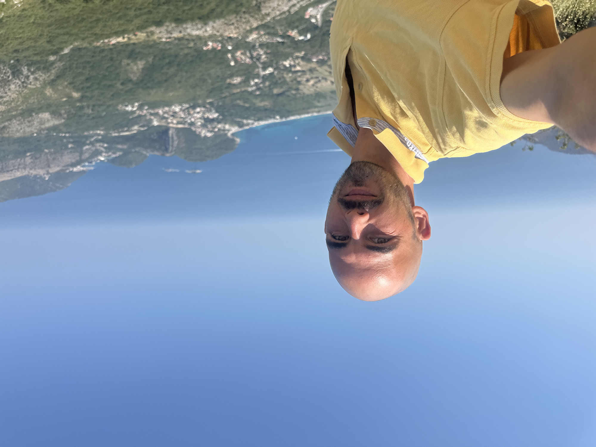 A man is taking a selfie outdoors with a clear blue sky and a coastal town in the background. The man has a shaved head, a mustache, and stubble. He is wearing a yellow polo shirt with a striped collar. The town appears to be nestled in hills overlooking the sea. The location is identified as Gluhi Do, Montenegro.