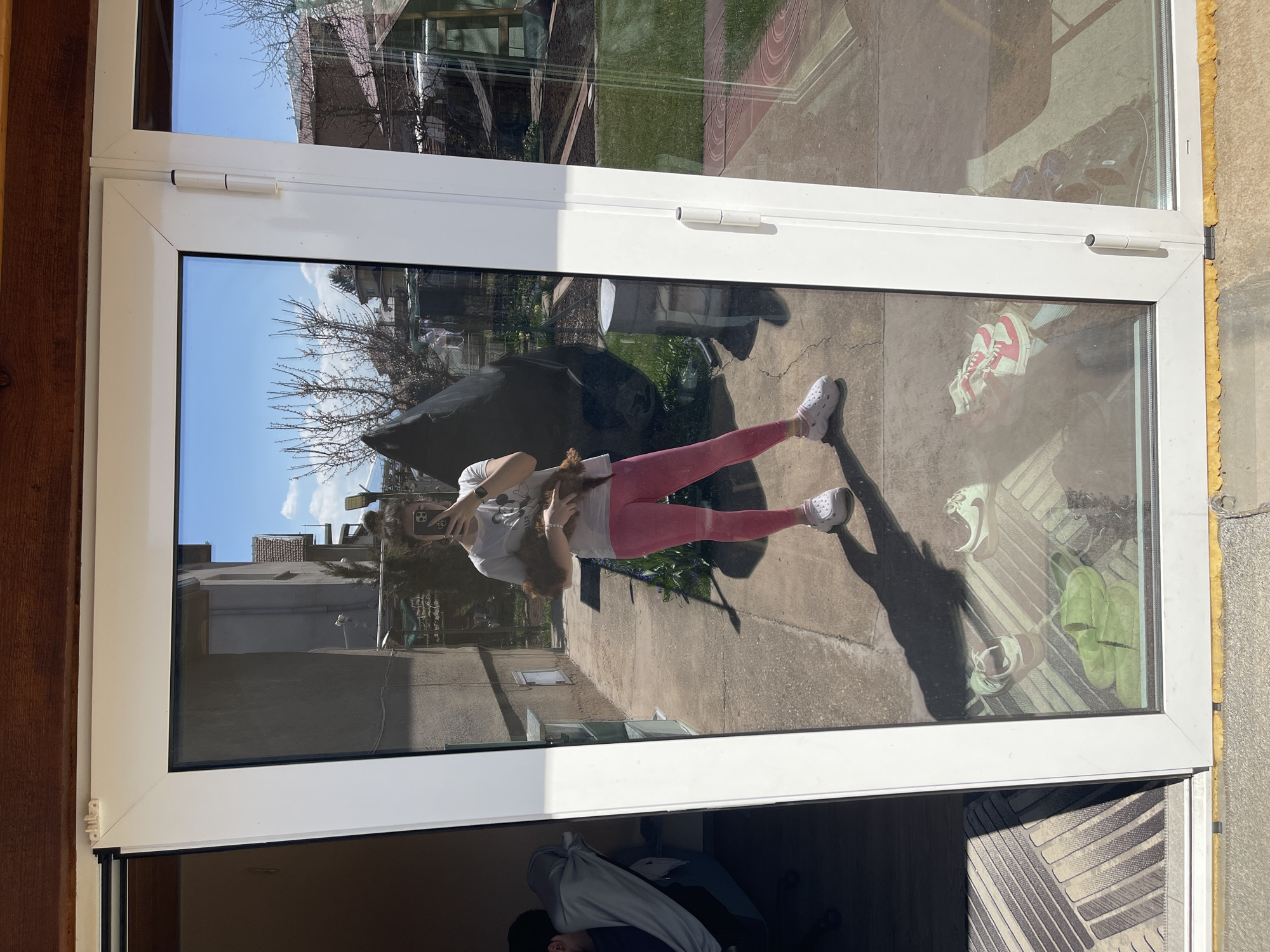 The image shows a reflection in a window or glass door of a person holding a small, brown dog. The person appears to be female, wearing pink leggings, white shoes, and a light-colored t-shirt. She is holding her phone, possibly taking a photo of the reflection. The background outside the window shows a sunny day with trees, buildings, and a gray concrete surface. A black tarp is visible in the reflection. Inside the building, other objects are visible, including shoes, a door mat, and possibly clothing. The image location appears to be in a residential area and likely located in the city of Sofia, Bulgaria. This assumption is based on information provided in the description prompt.