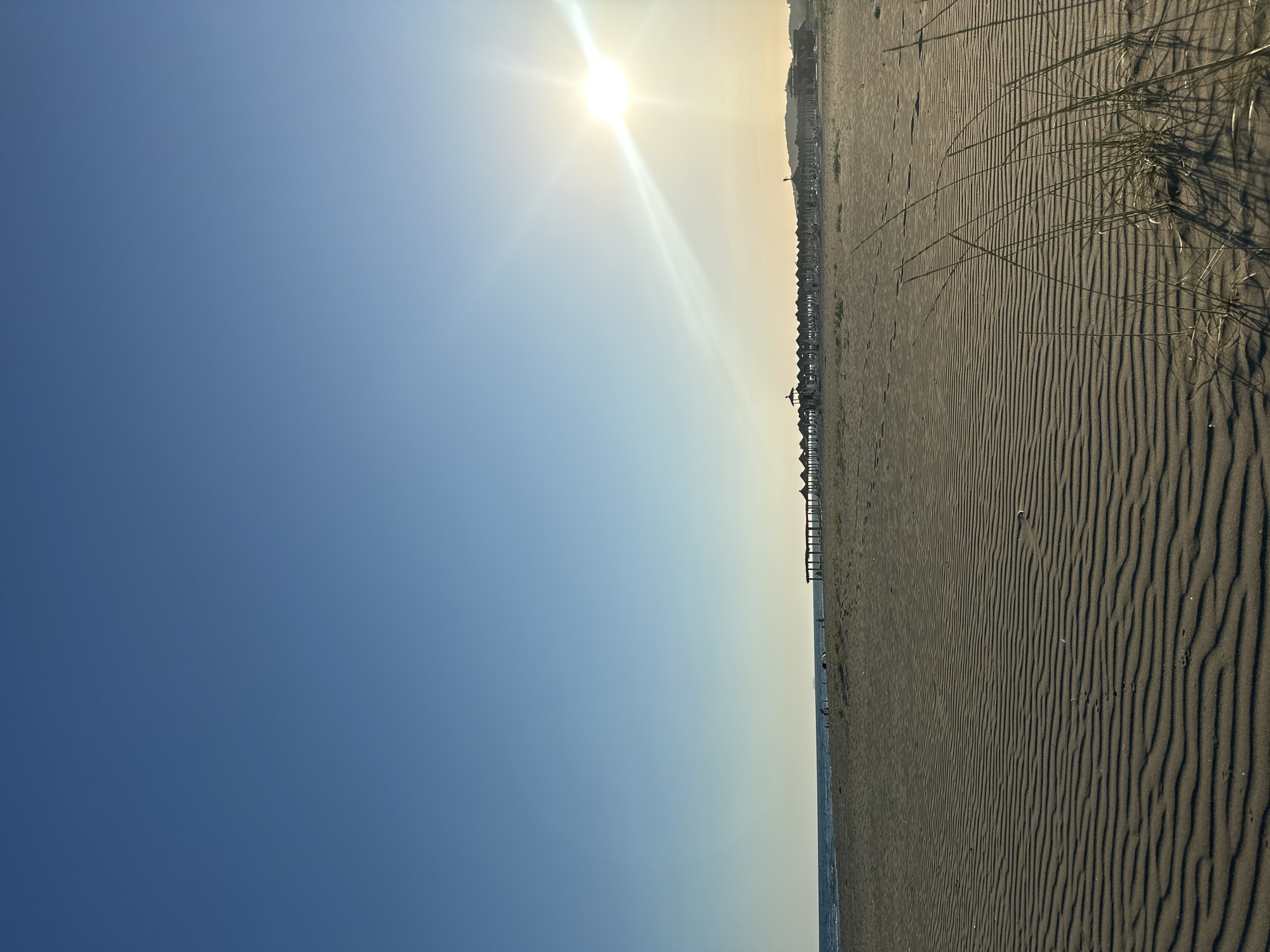 The image depicts a beach scene in Donji Štoj, Montenegro, bathed in the warm glow of the afternoon sun. The foreground is dominated by rippled sand, showcasing the natural patterns formed by the wind. A few blades of dry grass sprout from the sand in the lower right corner. In the mid-ground, the beach stretches towards the sea, which is calm and blue, meeting the horizon under a clear, pale blue sky. Along the coastline, a long line of thatched-roof umbrellas and lounge chairs suggests a resort or a well-maintained beach area. Several small figures, presumably beachgoers, are scattered along the shoreline, some near the water and others by the umbrellas. The sun, a brilliant orb with radiating rays, is positioned in the upper right quadrant of the sky, casting a bright glare. A gentle slope of land is visible in the far distance, against the backdrop of the clear sky.