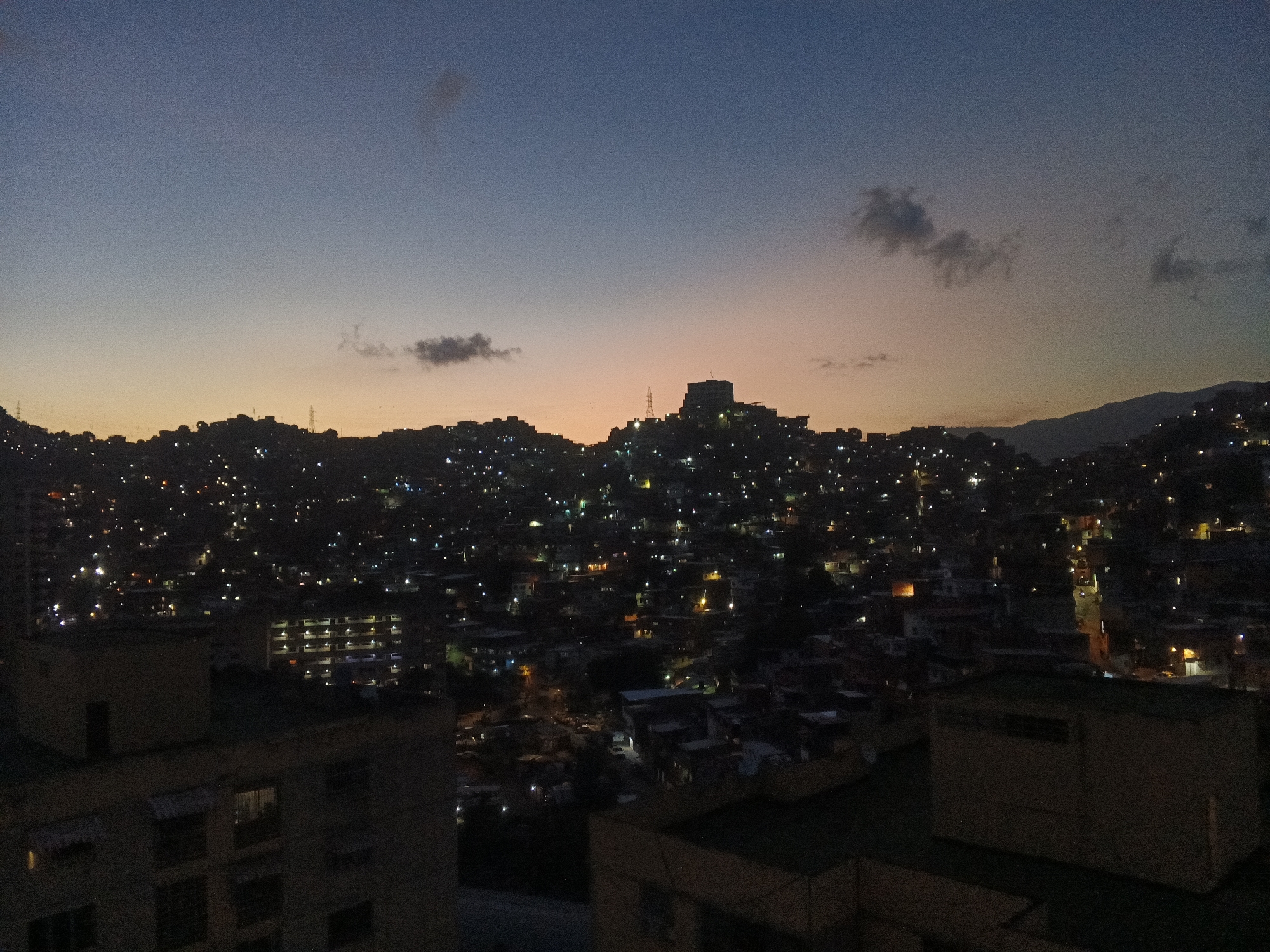 This is a high-angle, low-light photograph taken at dusk overlooking a densely populated hillside in Caracas, Venezuela. The sky is a gradient of deep blue at the top transitioning to pale orange near the horizon, with a few scattered clouds. The hillside is covered with countless small houses, many of which are illuminated by lights, creating a twinkling effect that stretches across the landscape. In the middle ground, a prominent building or structure with lights is visible on the highest point of the hill, possibly a lookout point or a significant edifice. Several power lines and transmission towers are also discernible, integrated into the urban sprawl. In the foreground, the dark silhouettes of apartment buildings or larger structures are visible, their windows unlit or dimly illuminated. The overall impression is of a vast, sprawling urban area at the foot of a mountain range, characteristic of the geography of Caracas, where informal settlements often climb the steep hillsides surrounding the city. There are no visible people or discernible actions taking place in the scene; the focus is on the cityscape and the transition from day to night.