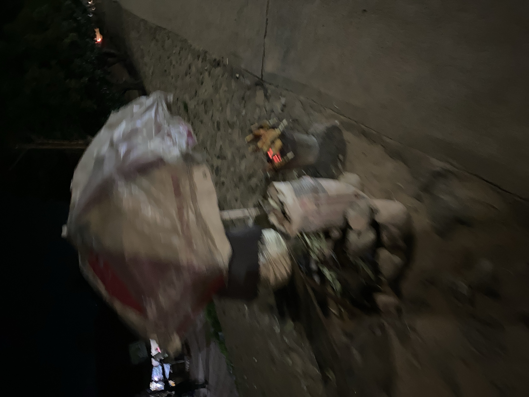 The image depicts a night scene in Bukuru, Nigeria, featuring a street food vending setup. A large umbrella, covered in plastic sheeting, is prominently displayed, suggesting it is protecting goods from the elements or for display. Beneath the umbrella, a food stall is visible with several items, including what appears to be corn being roasted over a small fire in a metal receptacle. The ground is uneven and appears to be a mix of dirt and debris. In the background, trees and streetlights indicate an urban or semi-urban environment. The overall impression is one of a humble, yet active, street vending operation at night.