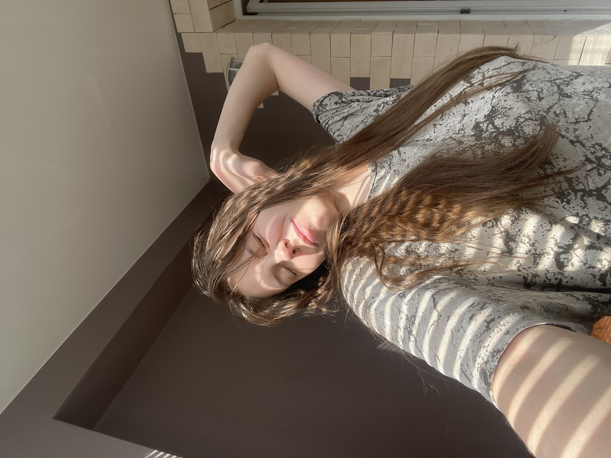 A young woman with long brown hair is lying down indoors. She is wearing a patterned gray t-shirt. Sunlight is shining on her and casting shadows. The location cannot be determined as being in the city of Ufa, Russia based on the given image.