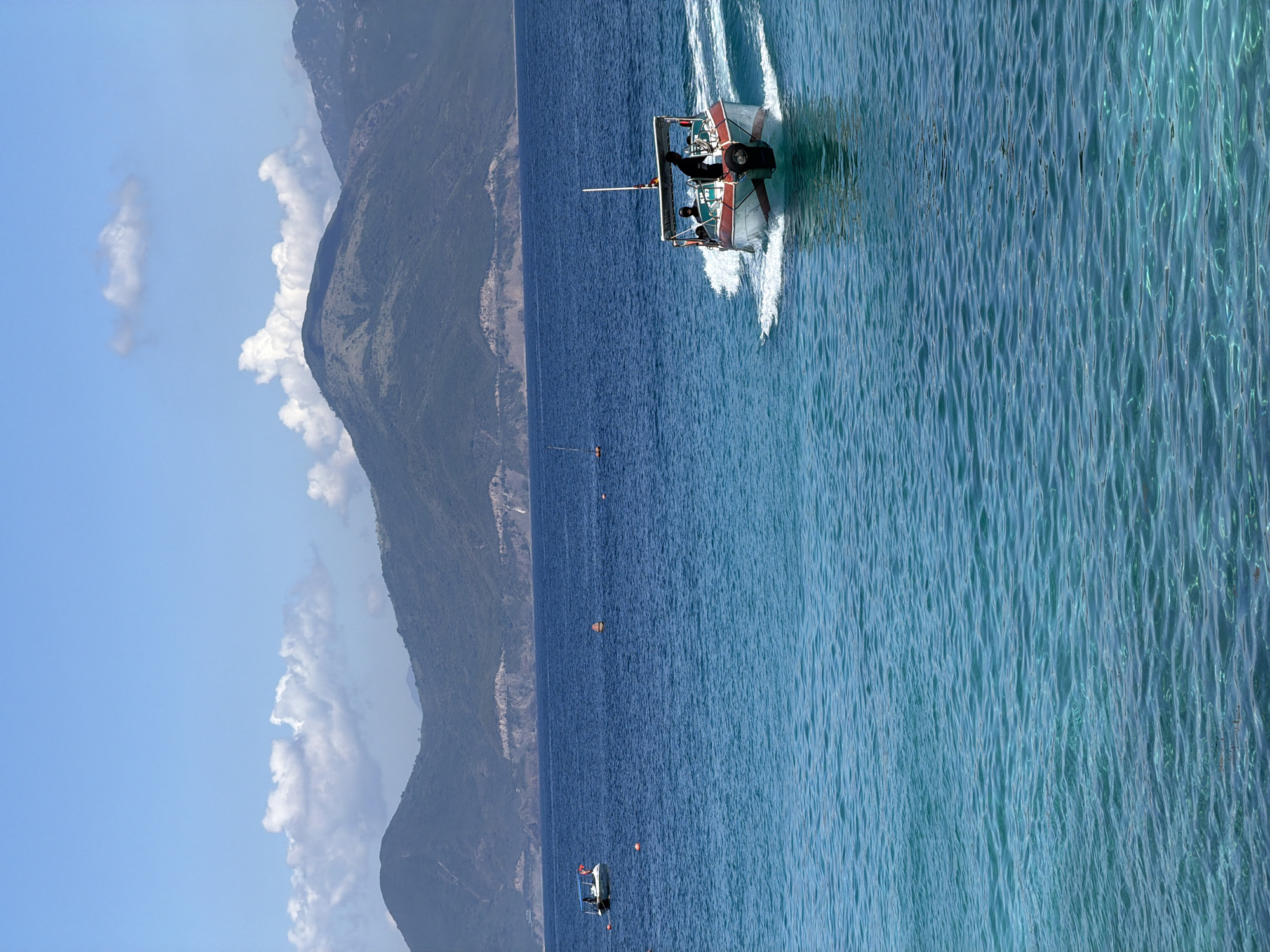 The image shows a body of water, likely the sea, with a speedboat moving through it. The water transitions from a darker blue in the deeper areas to a lighter, more turquoise color near the edge of the frame. The speedboat leaves a white wake behind it, indicating its movement. There are at least two people visible on the boat. On the left side of the frame, there is a coastal mountain range under a bright blue sky with scattered white clouds. In the water, there are some small orange buoys. Based on the description, the location may be near Nha Trang Ward, Vietnam, given its coastal features and mountainous landscape.