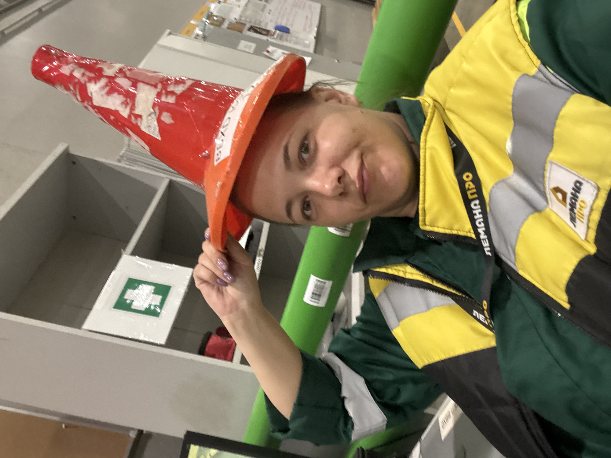 A woman wearing a green and yellow jacket and a red traffic cone on her head is looking at the camera with a slight smile. She is holding the cone with her left hand. The background includes a grey shelving unit with a first aid kit sign on one of the shelves and a green pipe or tube. The woman's jacket has text in Russian on the left side near the top.
There is nothing about this scene to suggest this is in the city of Lupanovo, Russia.